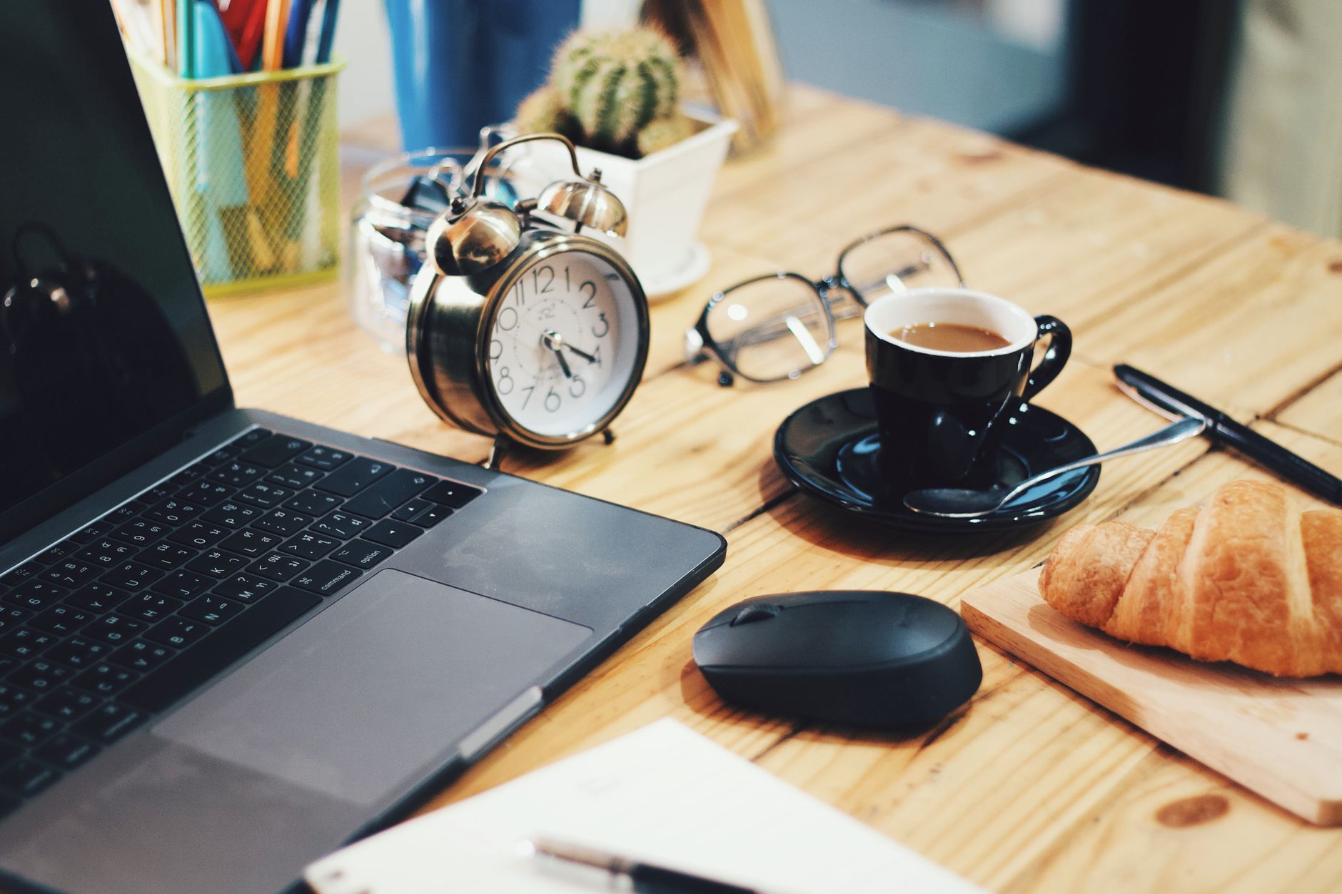 A warm workspace scene featuring a laptop, coffee, croissant, and clock on a wooden desk.