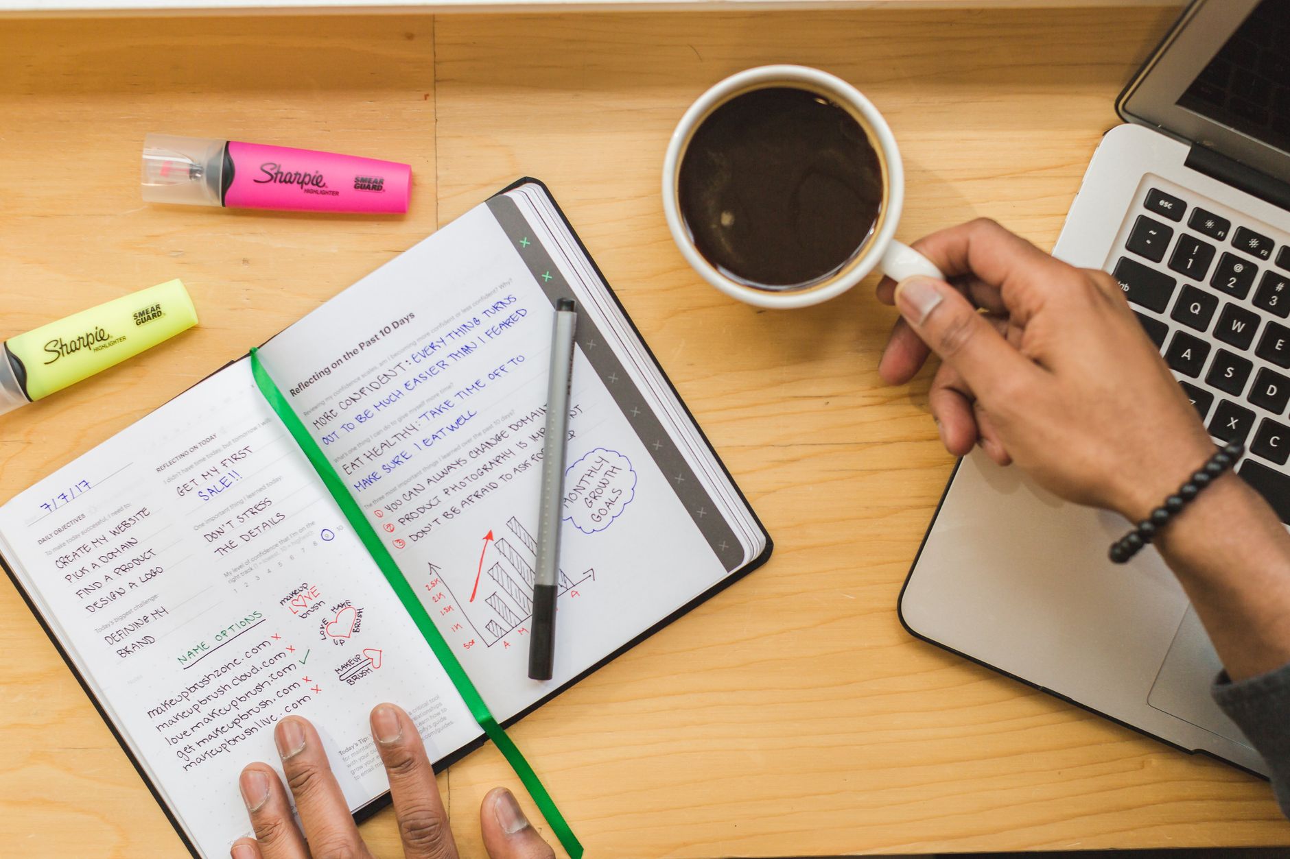 Top view of a person working with a notebook, coffee, and laptop on a wooden desk.