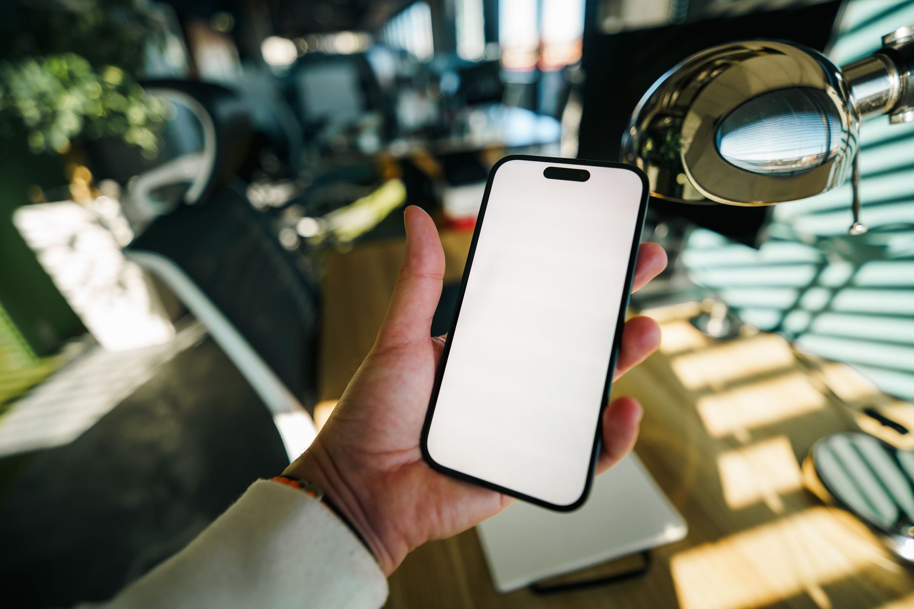 A hand holding a smartphone with a blank screen in a brightly lit modern office workspace.