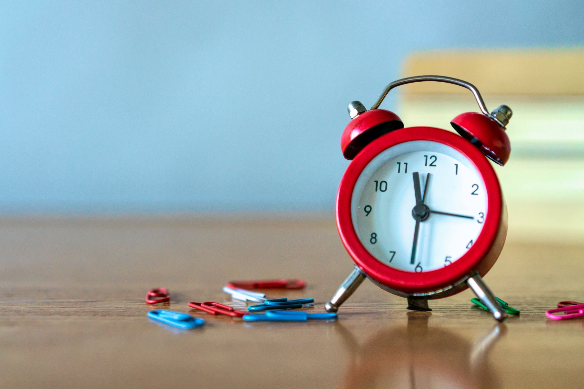 Red alarm clock and colorful paper clips on a wooden desk in a close-up view.