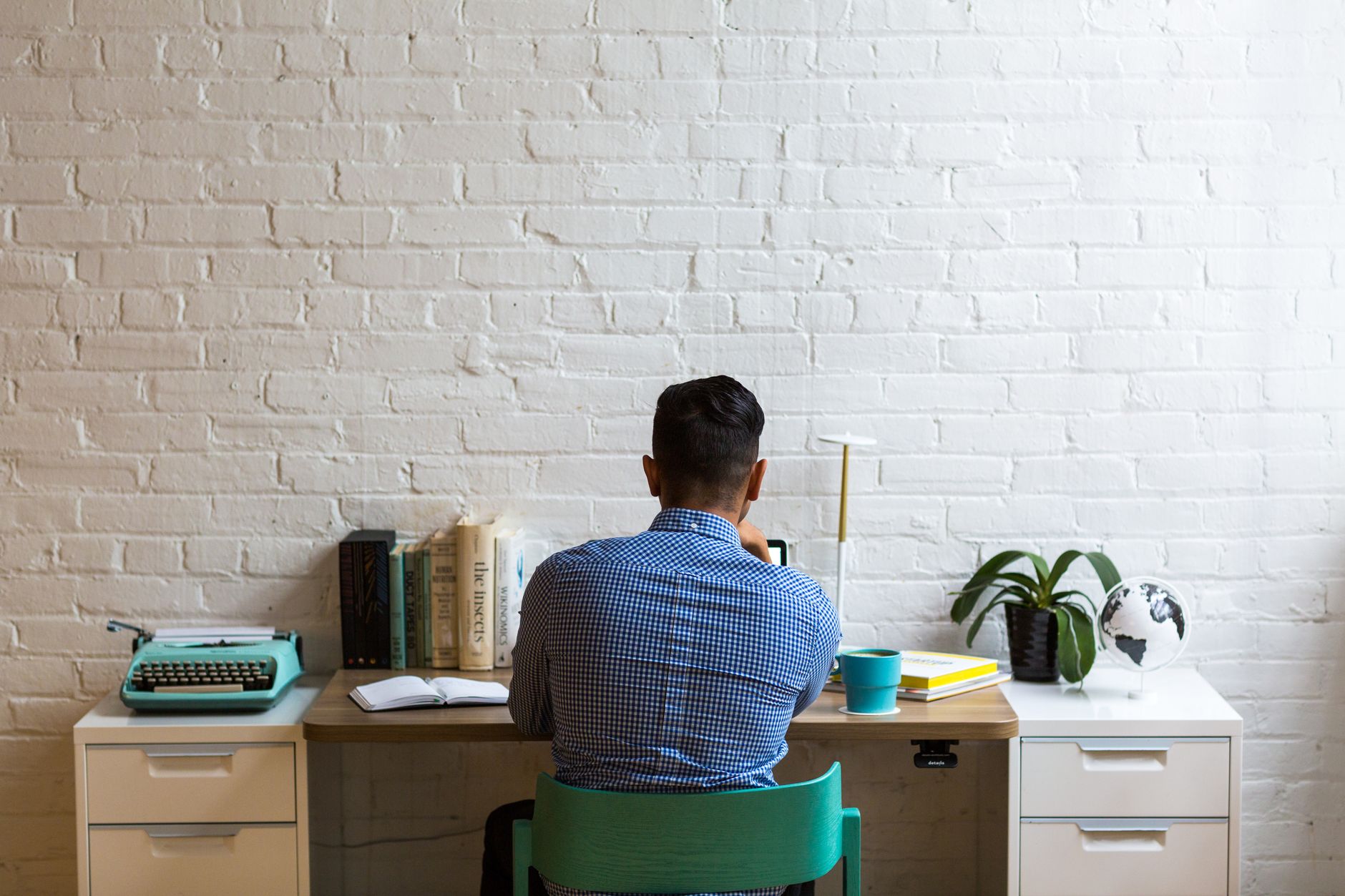 Back view of man at desk with typewriter and books, creating in a minimalist workspace.