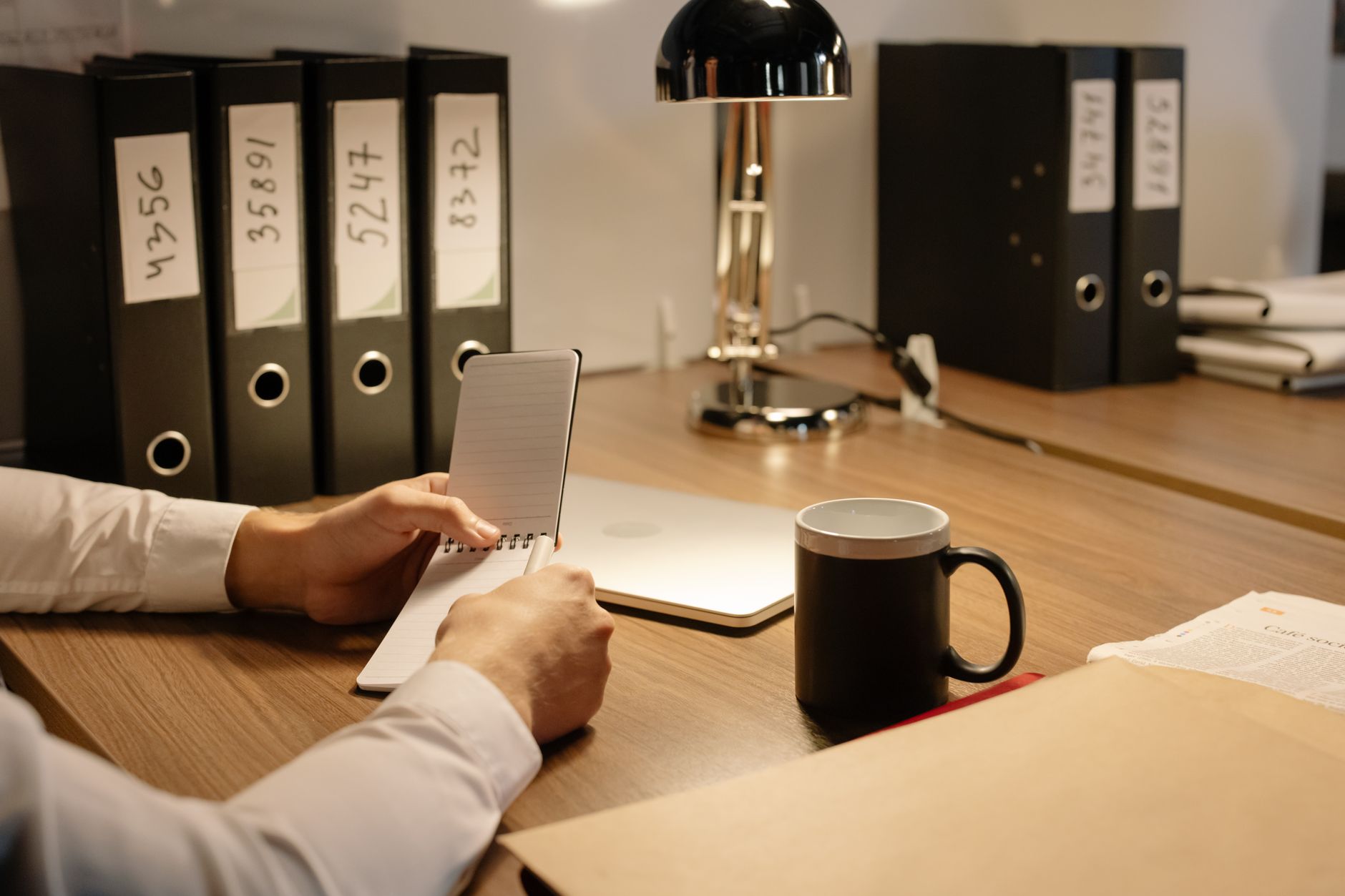 Office setting with a person holding a notebook, coffee mug, files, and laptop on a wooden table.