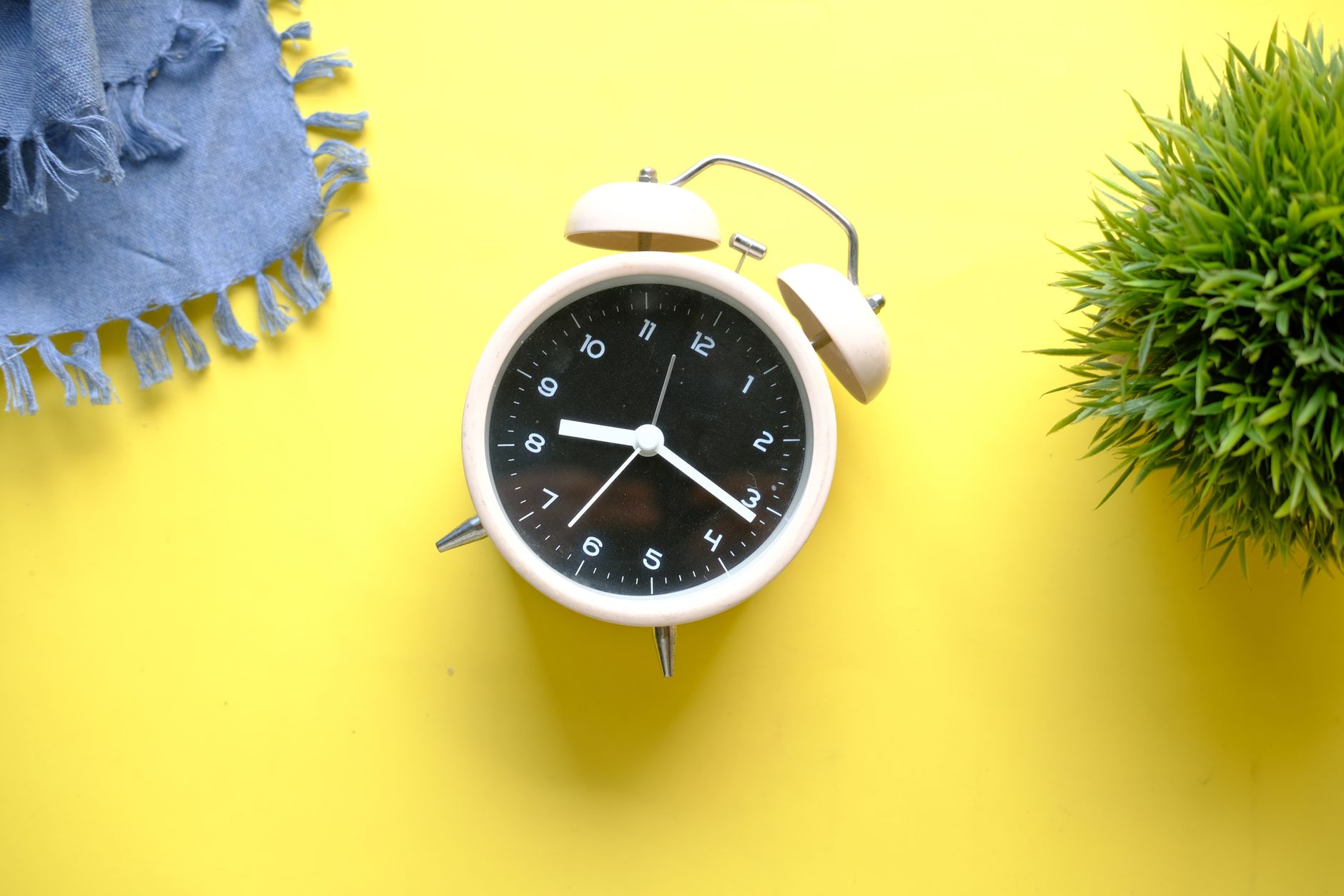 A vintage-style alarm clock on a vibrant yellow surface with a denim cloth and green plant.