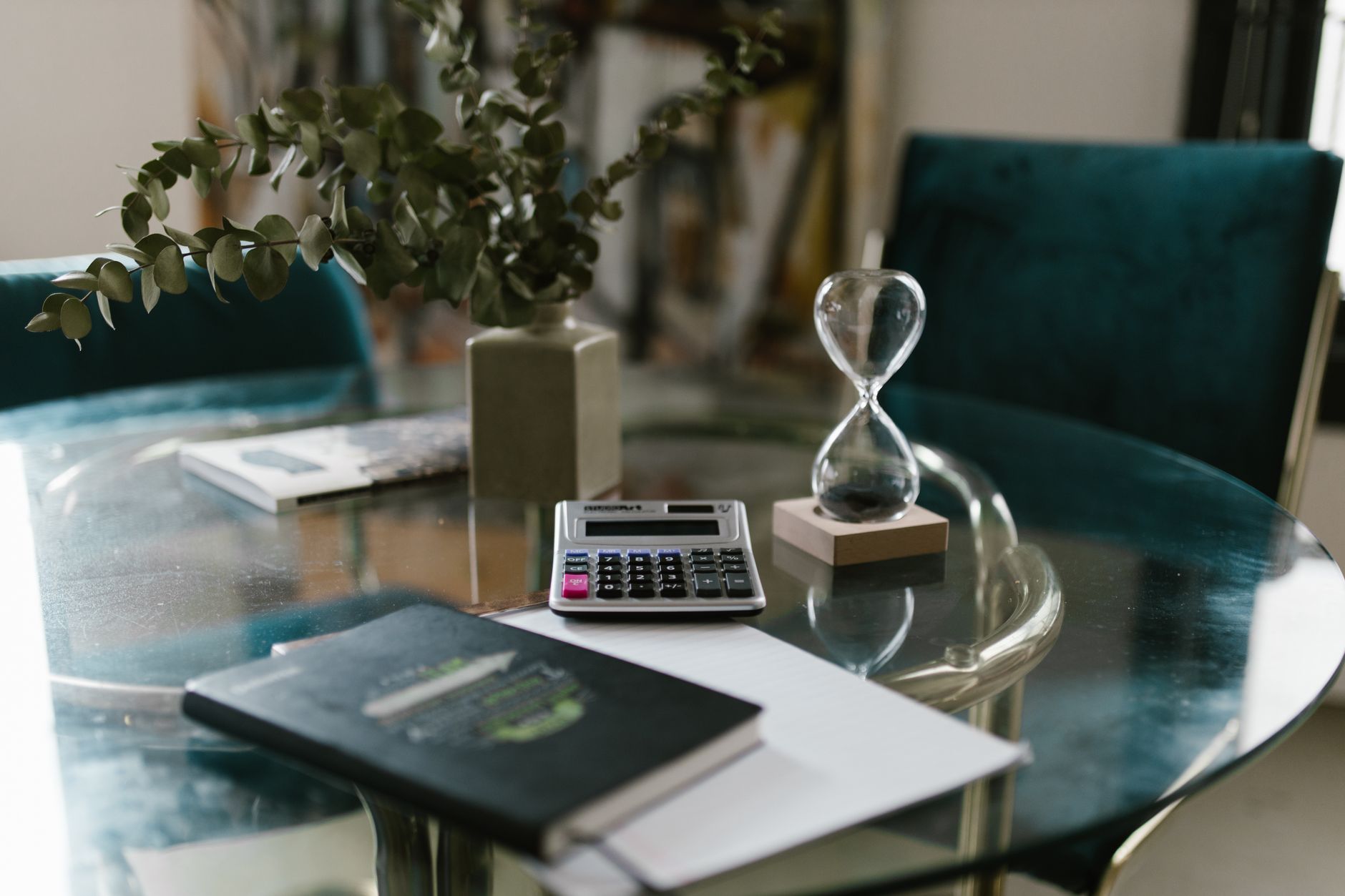 A minimalist workspace featuring a calculator, notebook, and hourglass on a glass table.