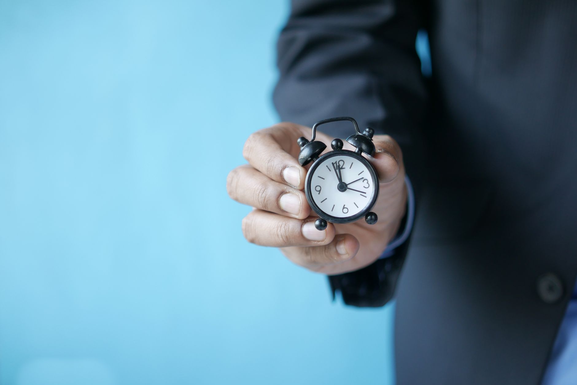 Close-up of a person's hand holding a small black analogue alarm clock against a blue background.