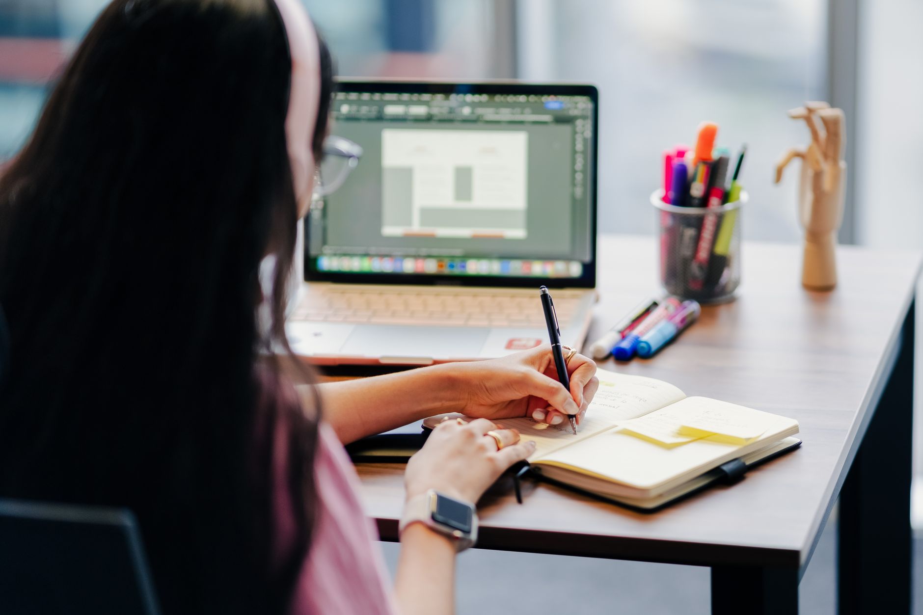 A woman multitasking by writing notes and using a laptop at a desk with colorful stationery.