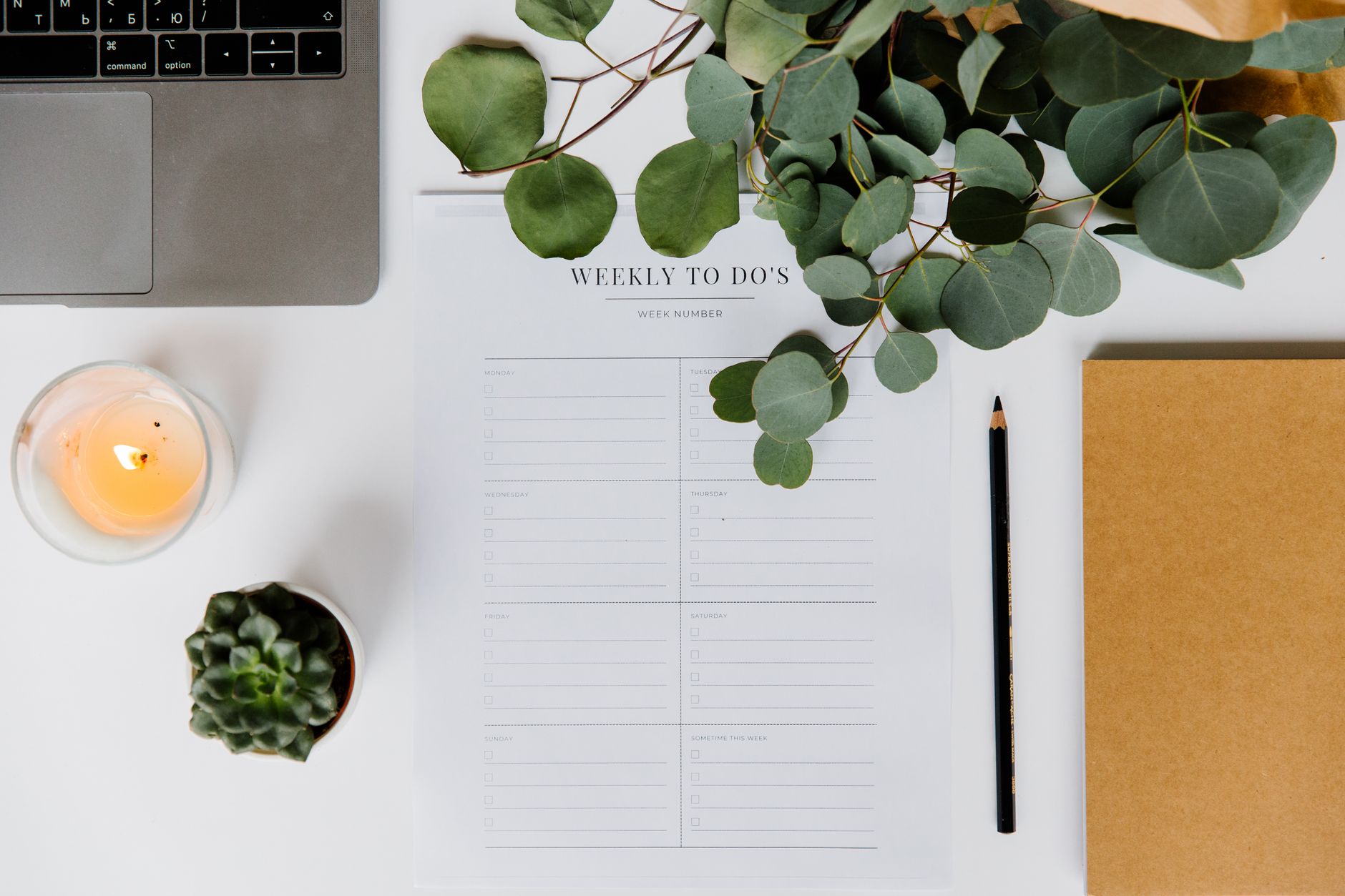 Flat lay of a well-organized workspace featuring a laptop, candle, eucalyptus leaves, and to-do list.
