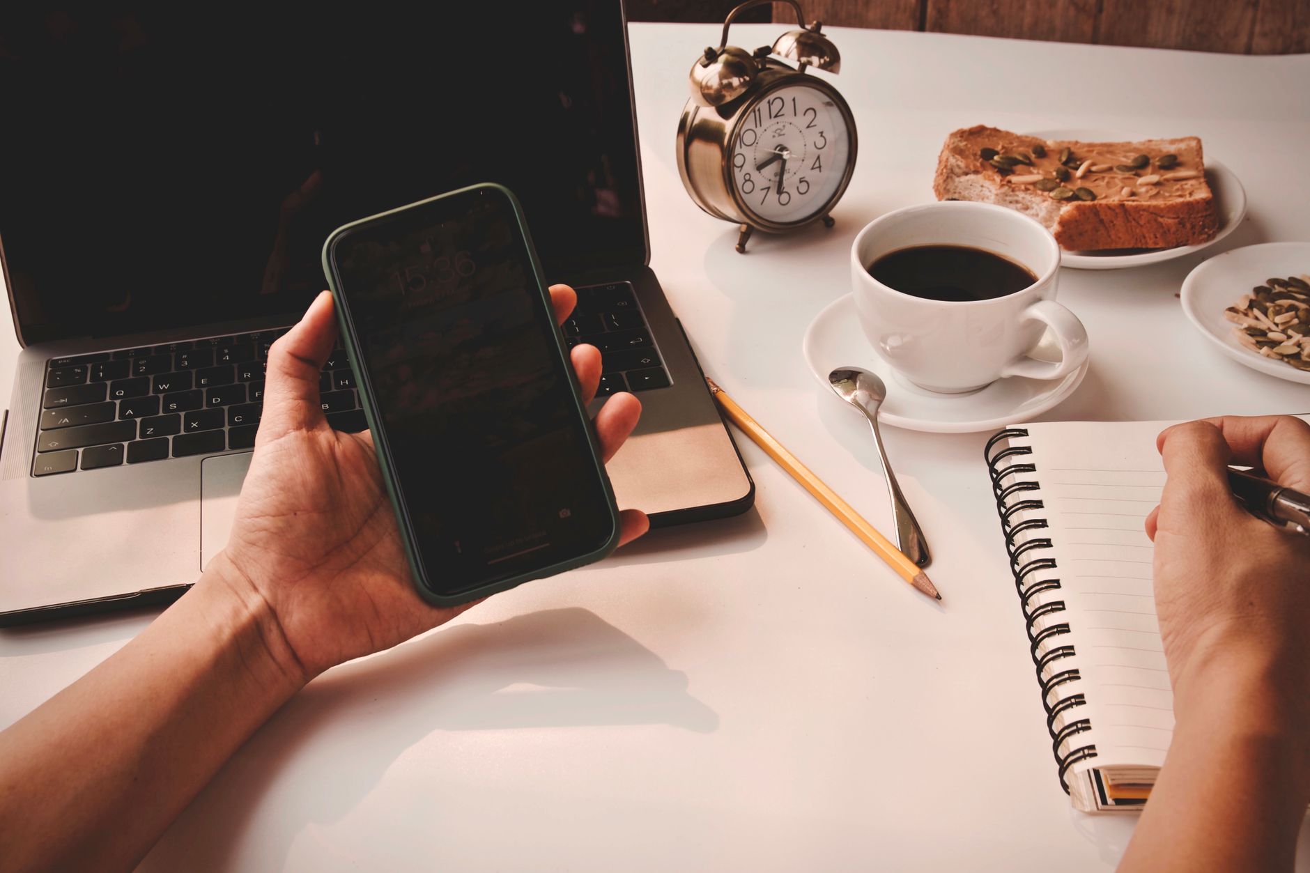 A cozy morning workspace with coffee, bread, laptop, and smartphone on a table.