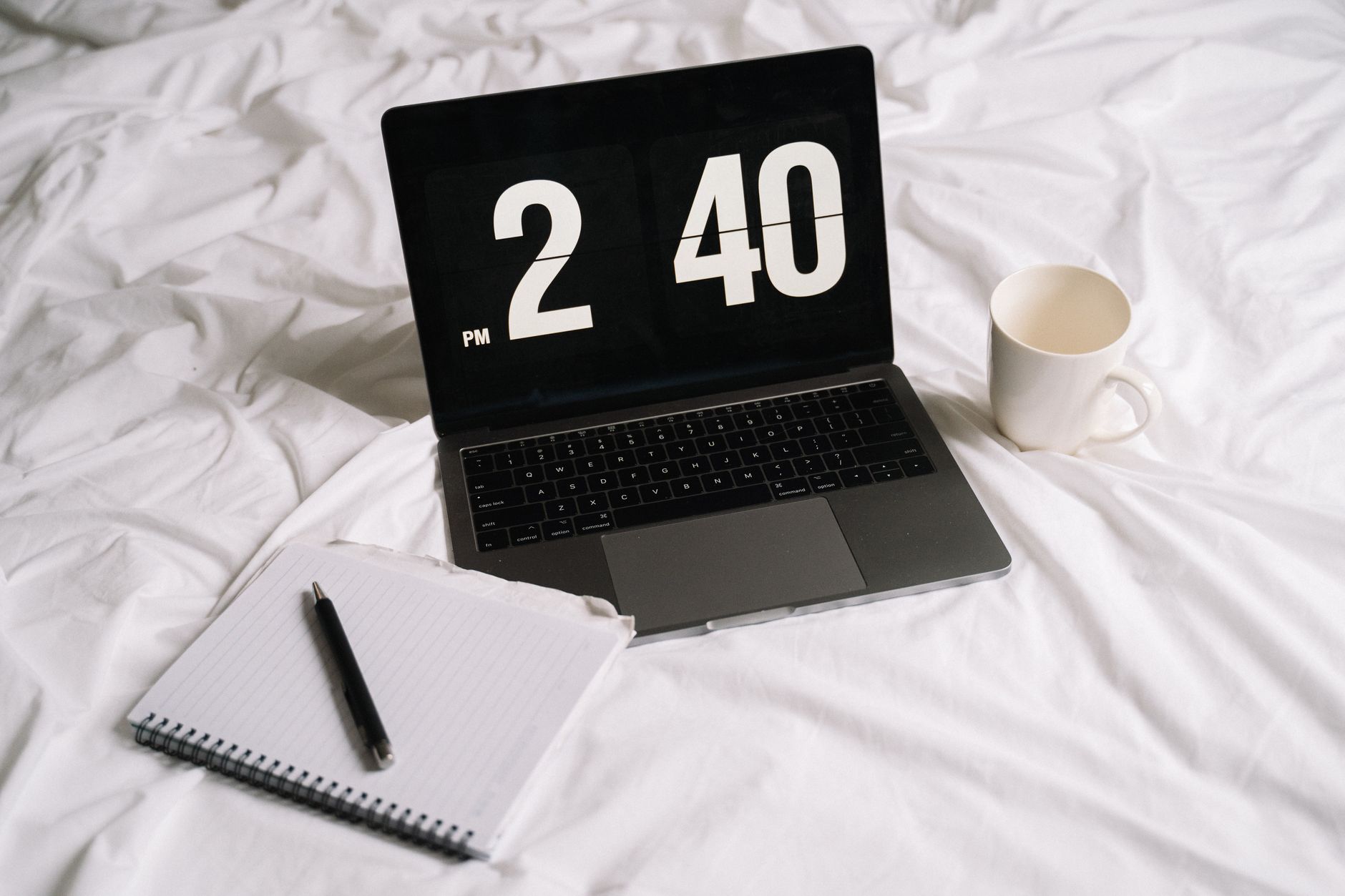 Laptop displaying time next to a notebook and coffee on a bed, creating a cozy work setup.