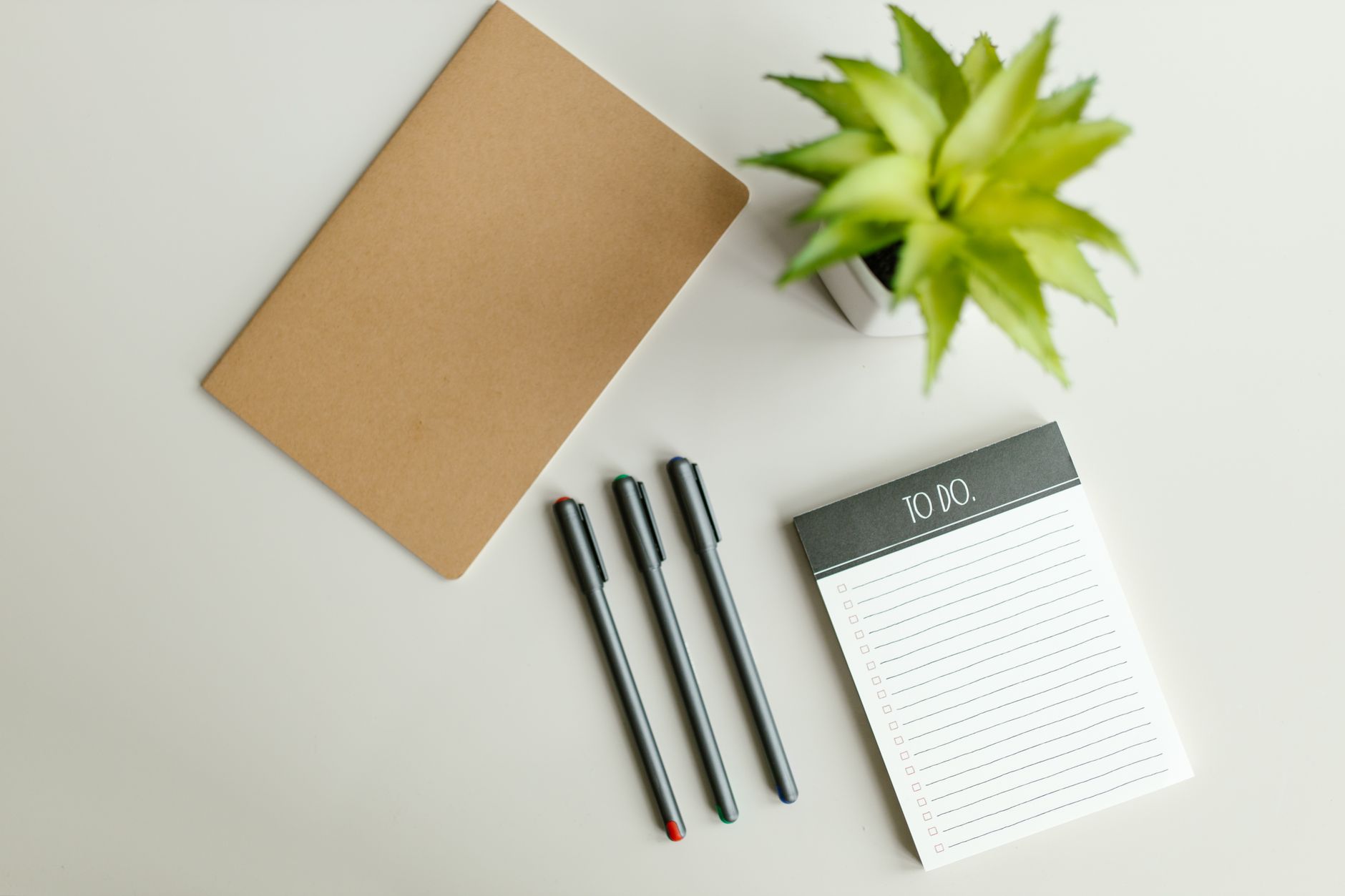 Flat lay of a to-do list, notebook, pens, and succulent on a light gray desk surface.