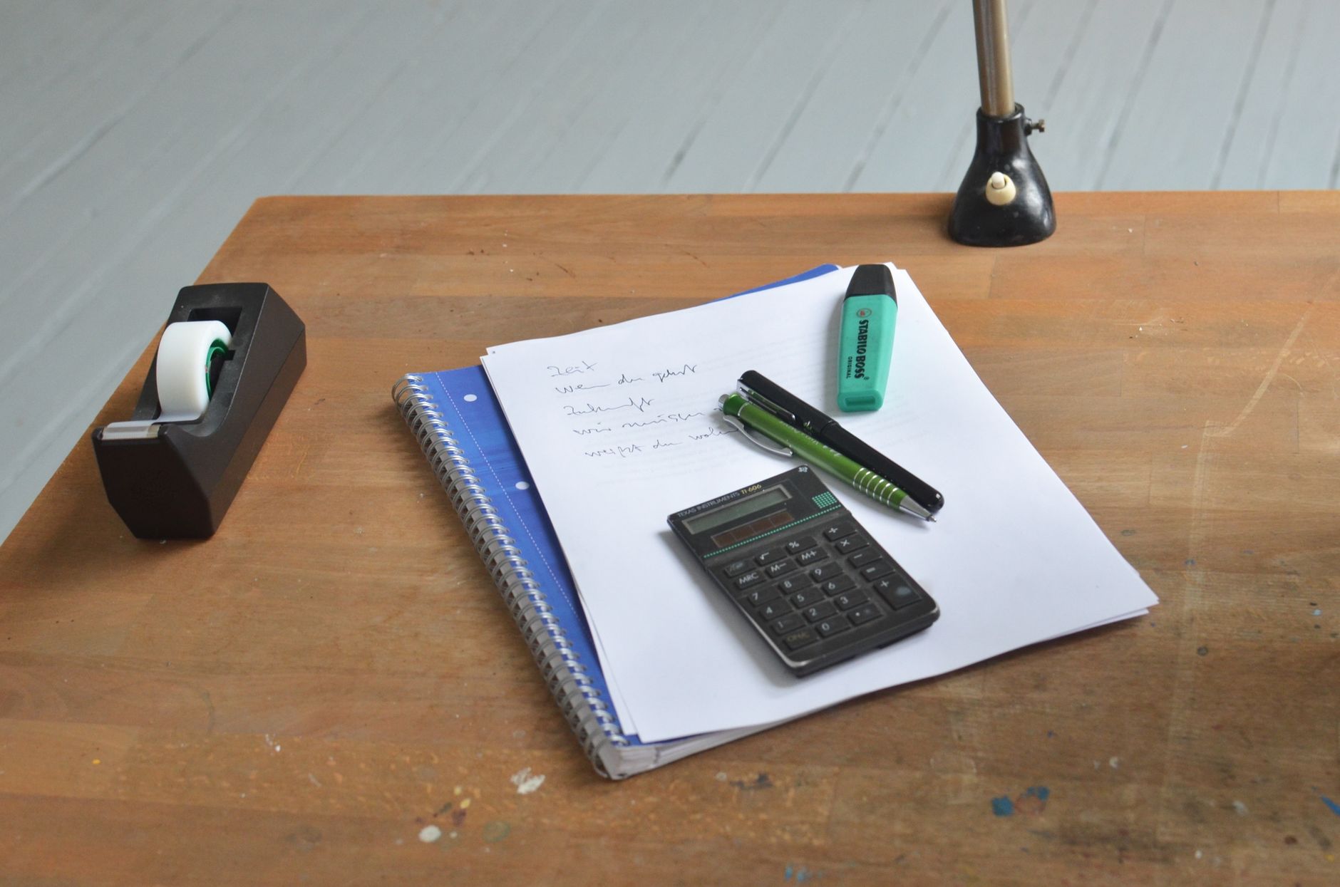 High angle of wooden desk with papers and small calculator with and pens on top near tape dispenser