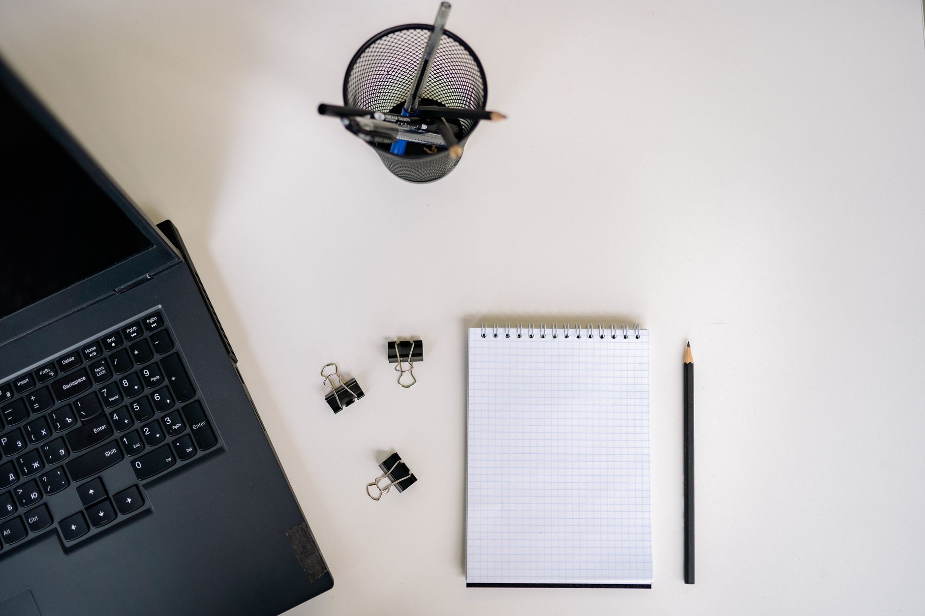 Top view of an office setup with a laptop, notepad, pencils, and clips on a white desk.