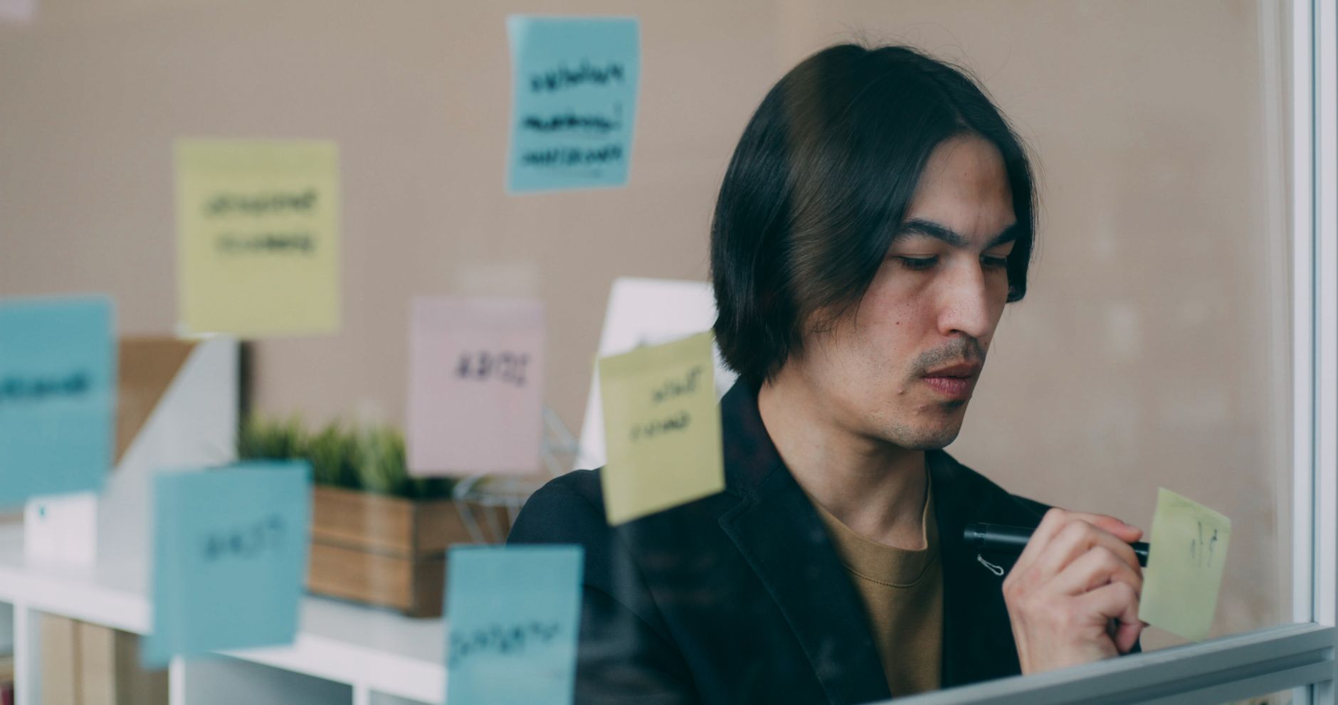 Focused young adult writing on sticky notes on a glass wall in an office setting.
