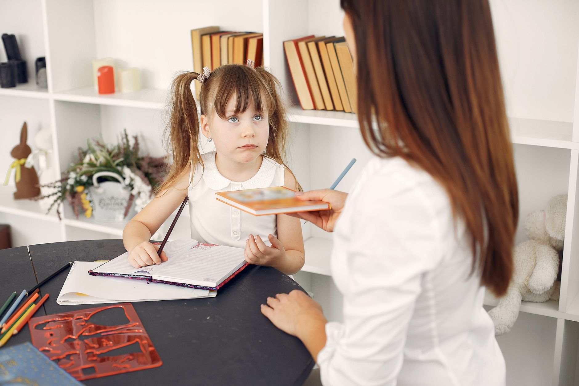 Little girl sitting at table writing in notebook with pencil while taking textbook from young female home teacher in white shirt during home education against shelf with textbooks