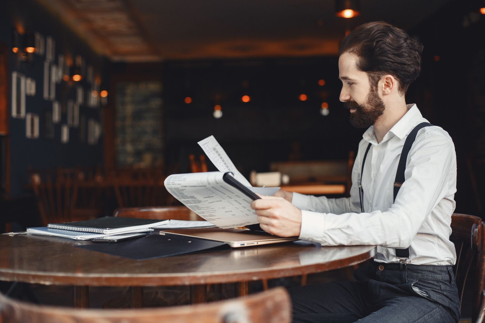 A man with a beard reads a printout while sitting at a wooden table in a cozy café setting.