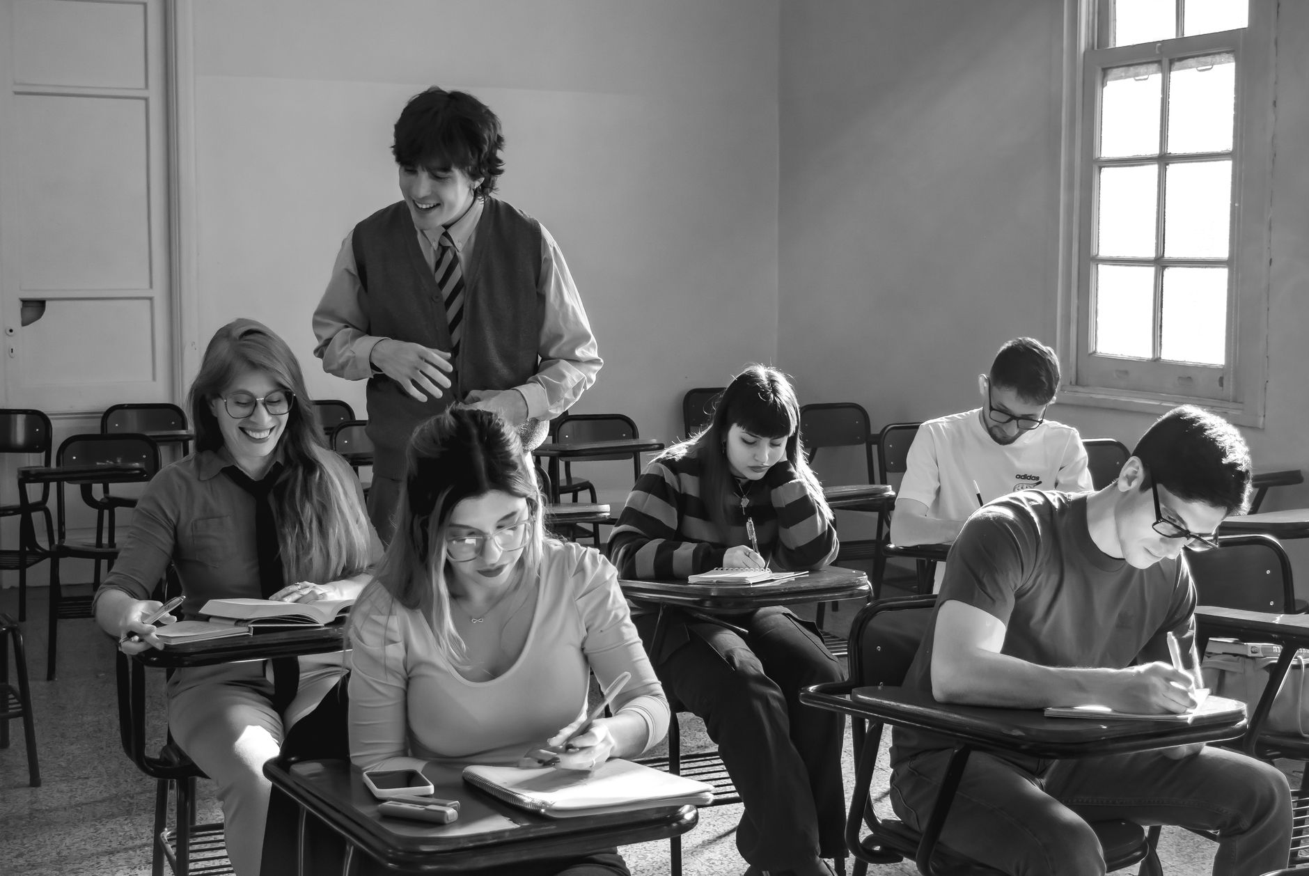 Black and white image of students writing in class while teacher observes.