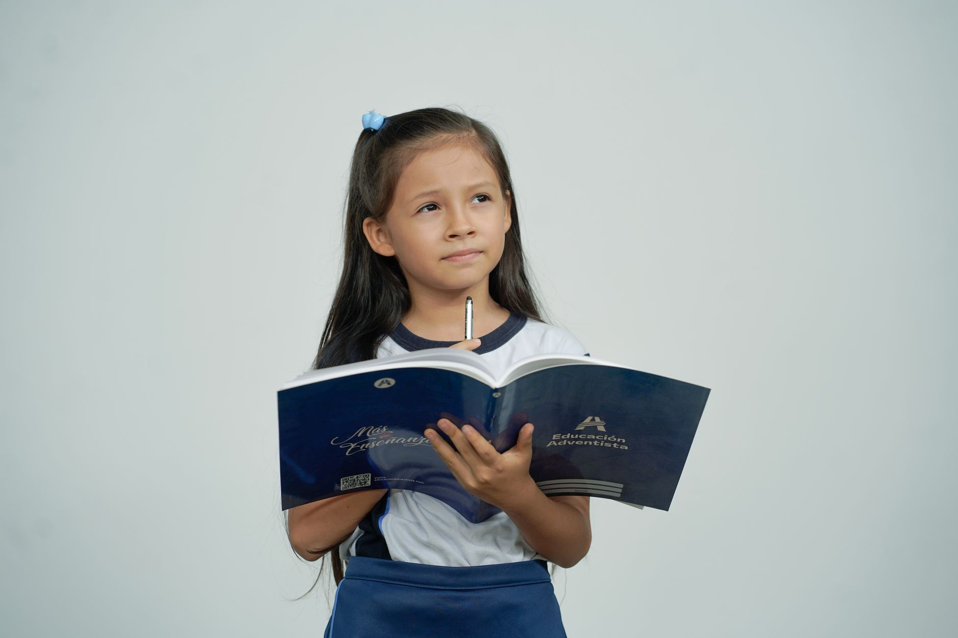 Young schoolgirl with long hair holding a book and pencil, deep in thought against a plain background.