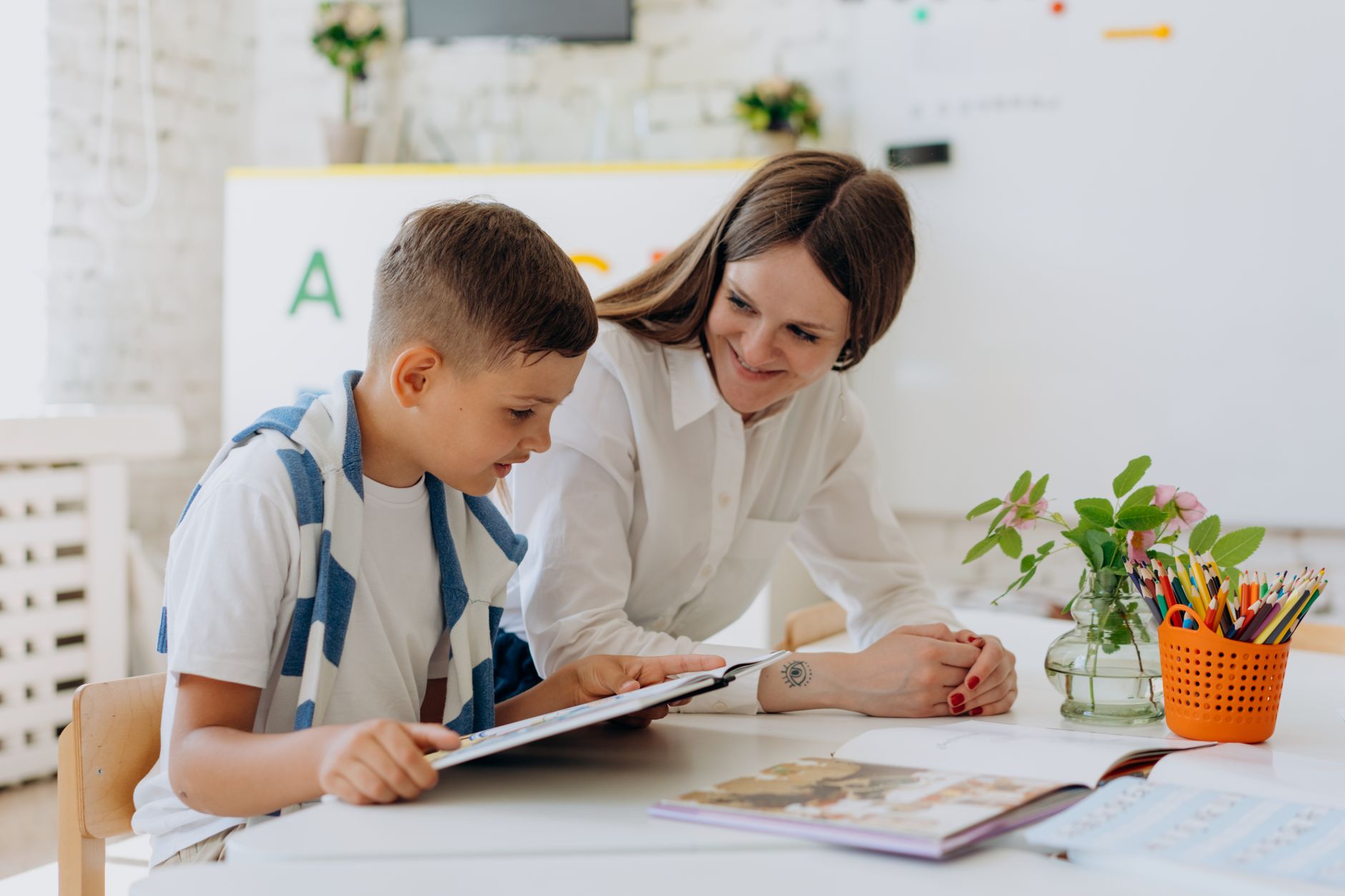 A teacher helping a young boy learn using a tablet in a bright classroom setting.