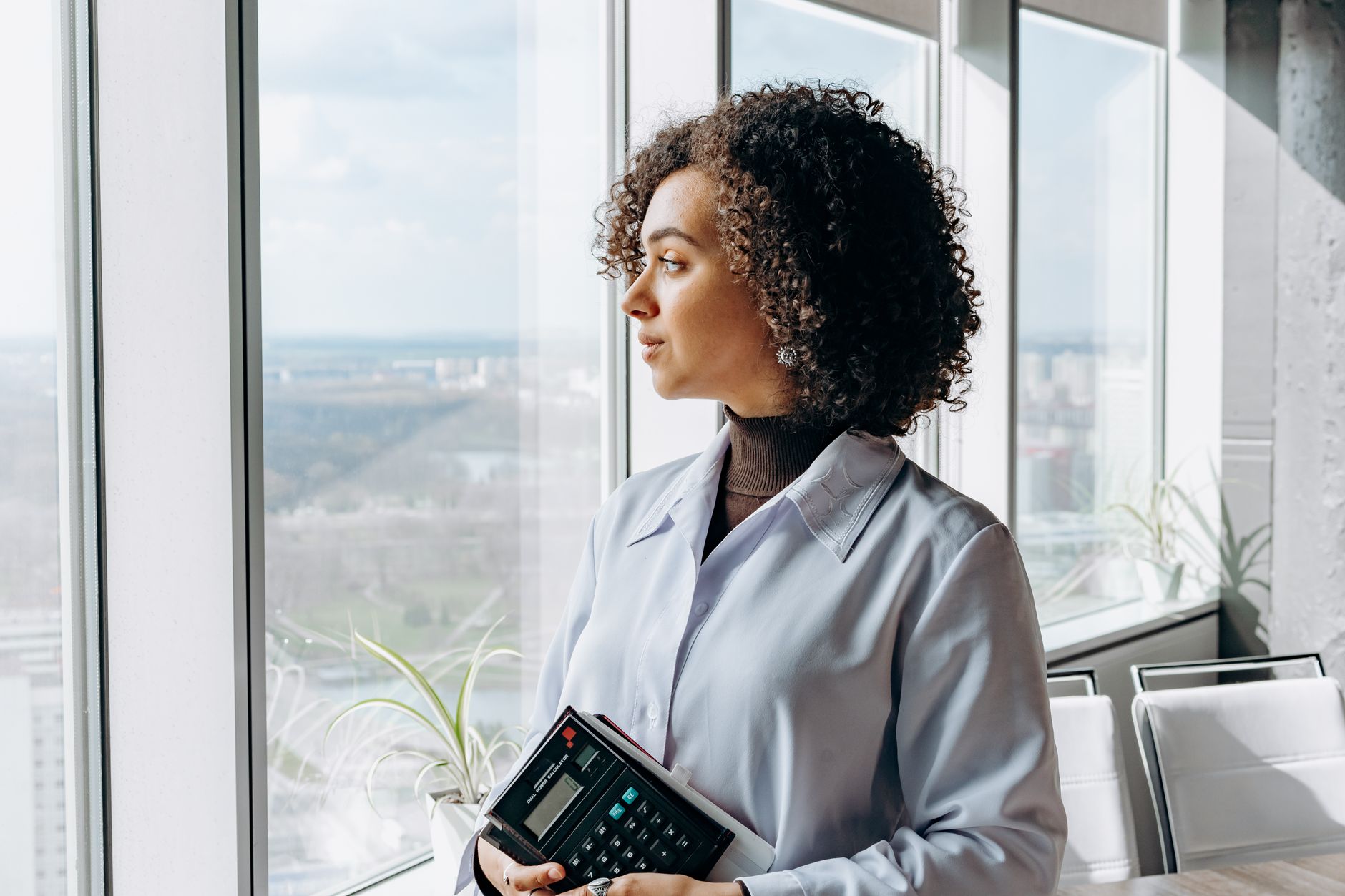Woman in office attire holding a calculator, gazing out a large window, with a cityscape view.