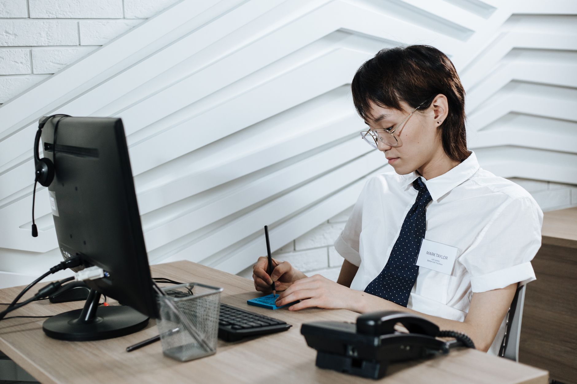 Asian call center agent writing notes at a modern desk setup with computer and headset.