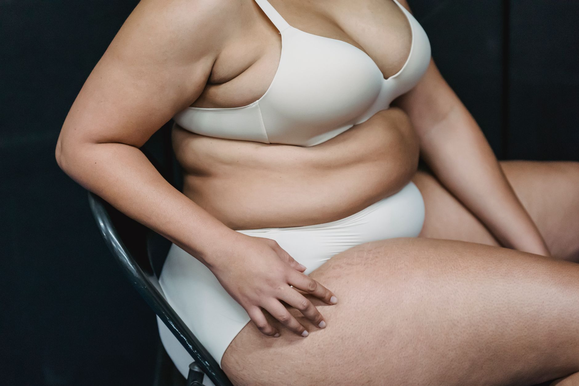 Crop anonymous plump woman in white underwear sitting on chair in light studio on dark background