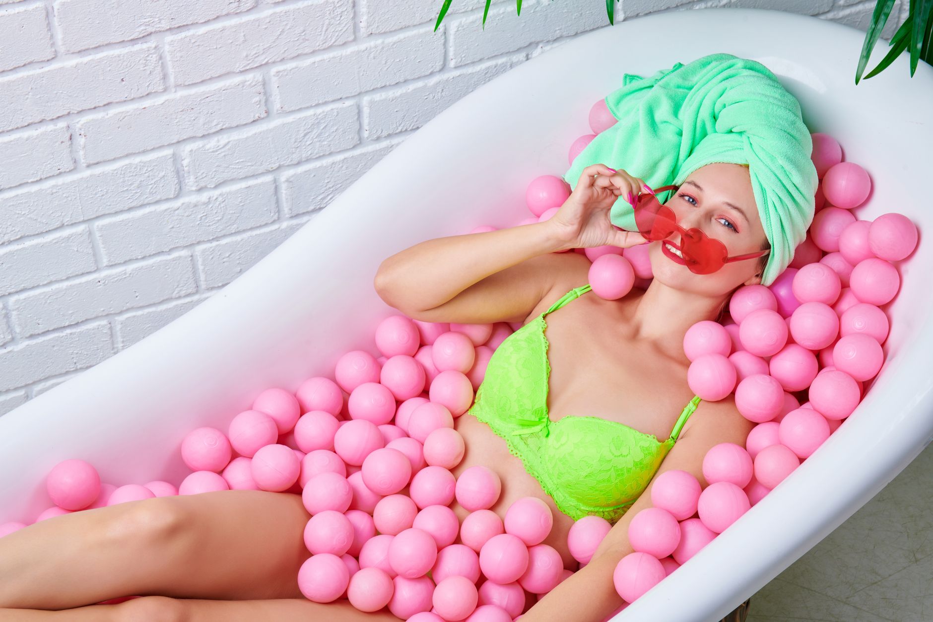 Woman in a bright setting relaxes in a bathtub filled with pink balls, wearing sunglasses and green lingerie.