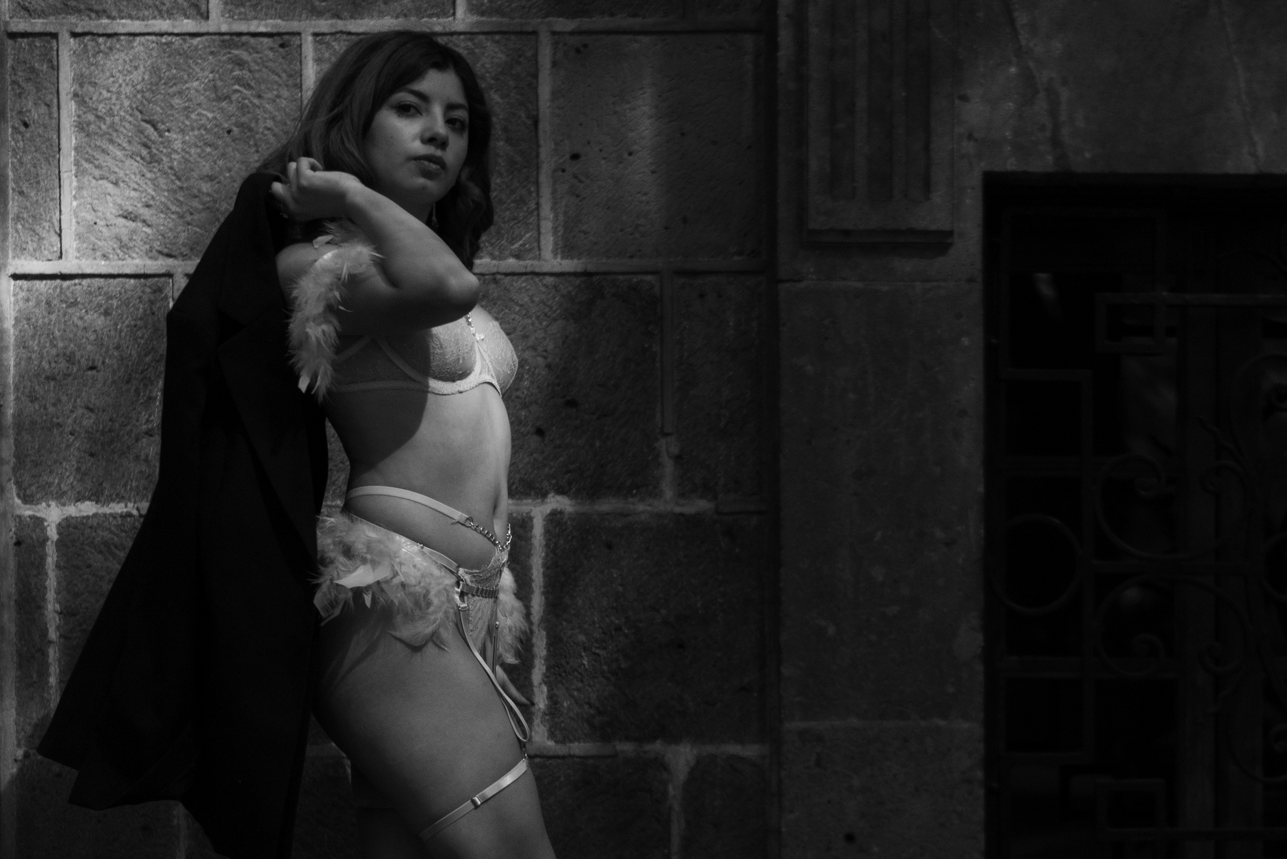 Elegant black and white portrait of a model posing in lingerie with a coat, set against a stone wall.