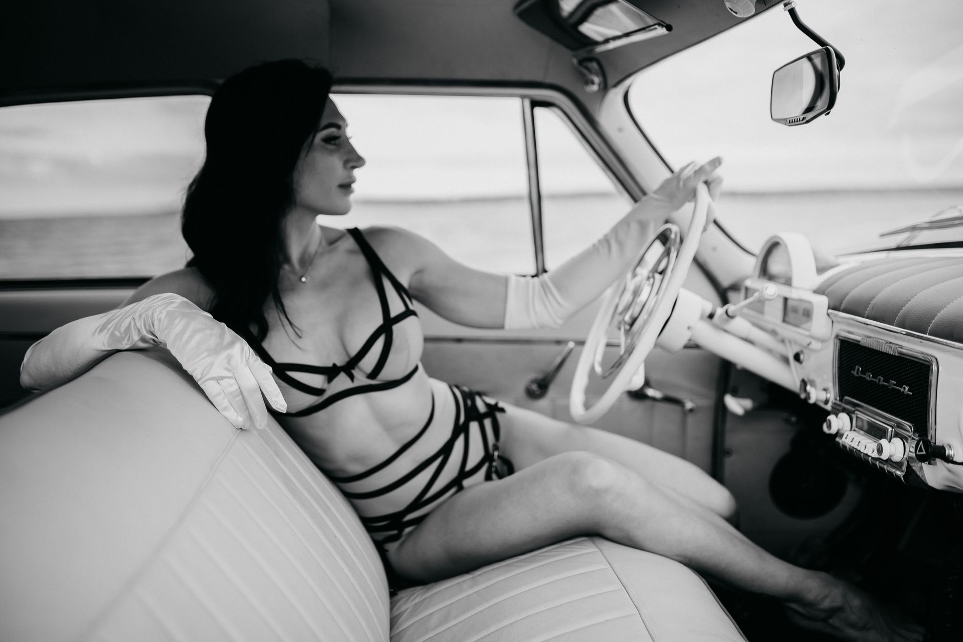 A beautiful woman in lingerie sits in a vintage car, presenting elegance and retro style.