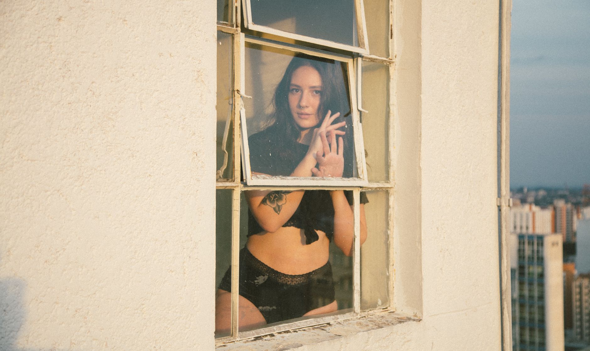 Artistic shot of woman posing in lingerie through a glass window, cityscape backdrop.
