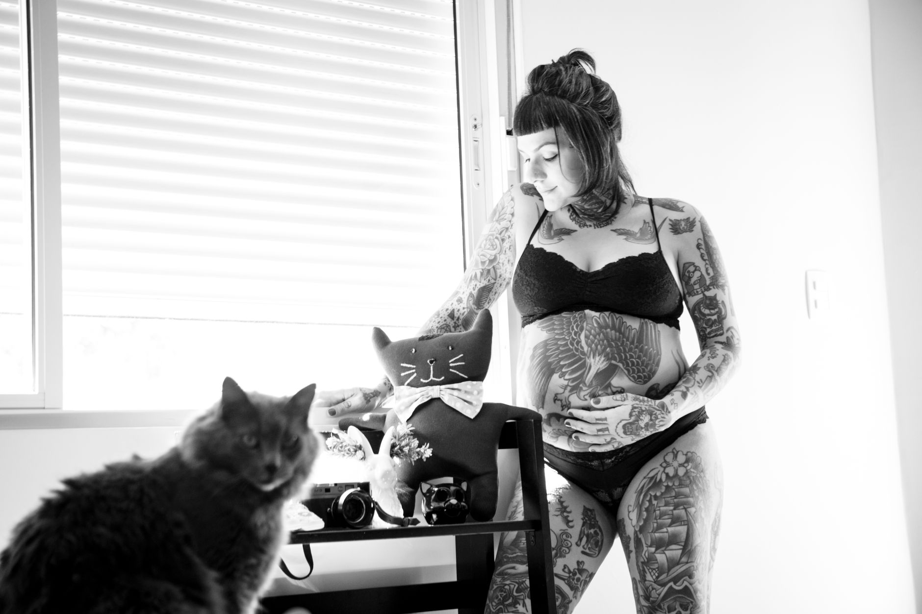 Intriguing black and white photo of a tattooed woman with a baby bump indoors with a cat.