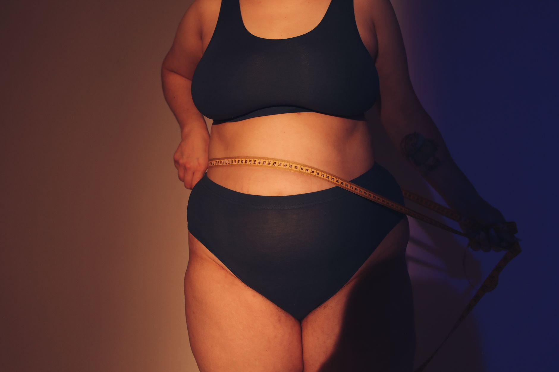 A body positive woman stands in a studio setting, holding a tape measure around her waist.