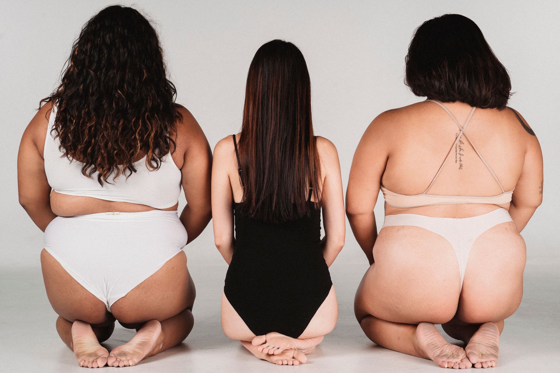 Back view of unrecognizable skinny woman in black bodysuit sitting on knees between overweight models in underwear on white background