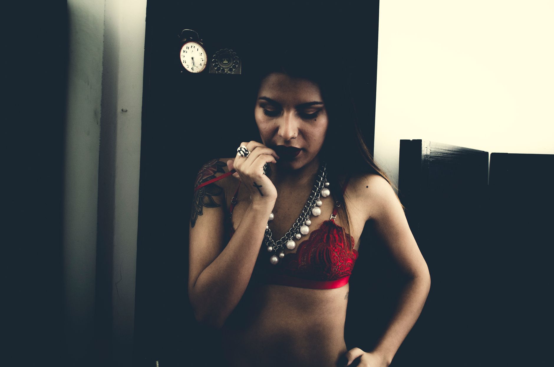 A woman in red lingerie, accessorized with a statement necklace, poses thoughtfully indoors.