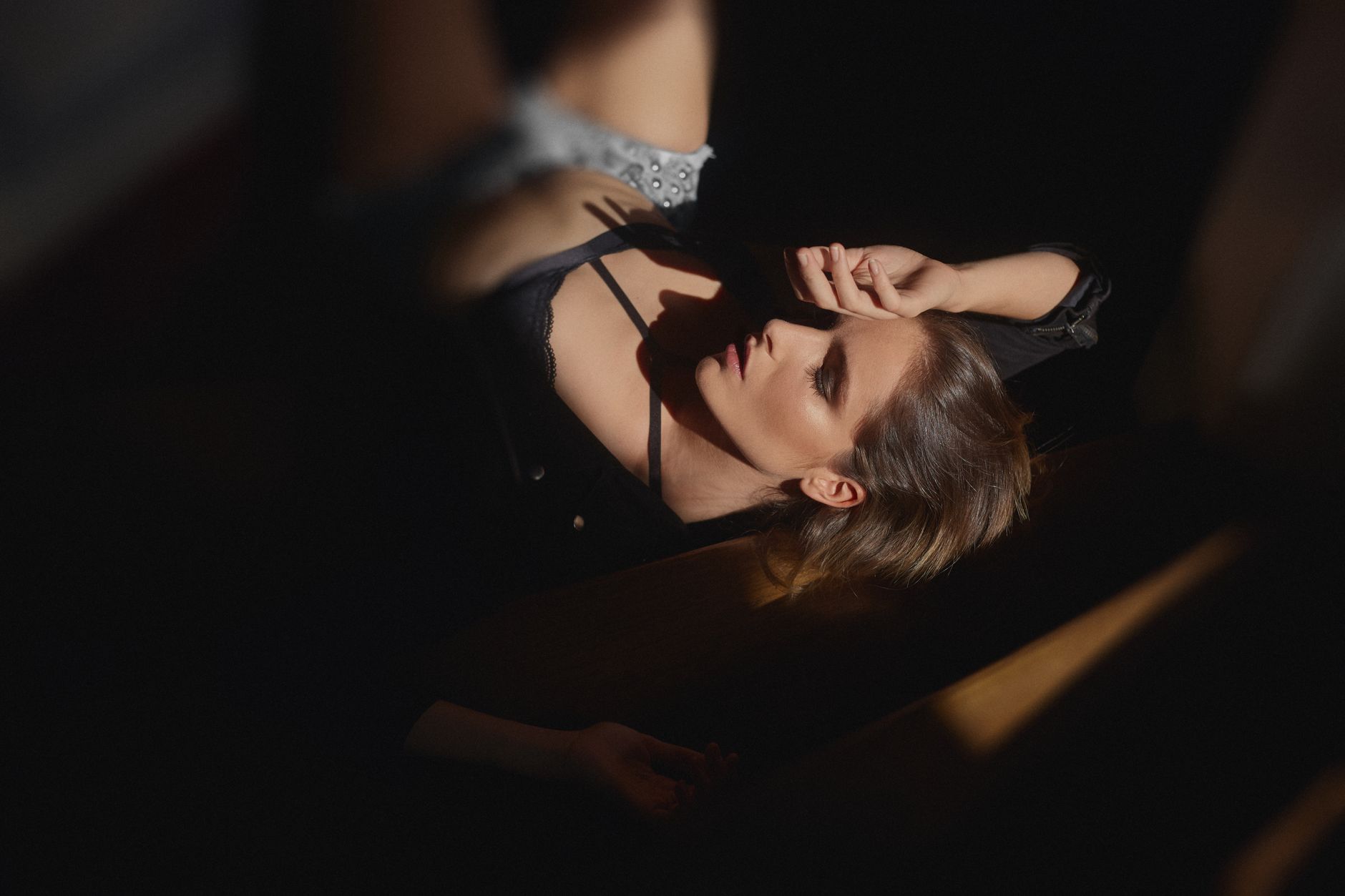 From above of young sensual female in black underwear touching face while lying in dark room with closed eyes