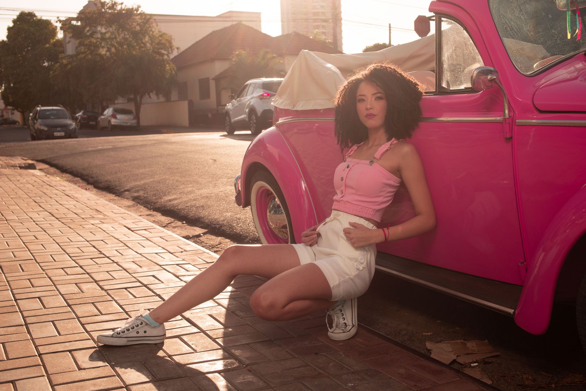 Stylish young woman in pink top and white shorts posing by a vibrant pink classic car on a sunlit street.