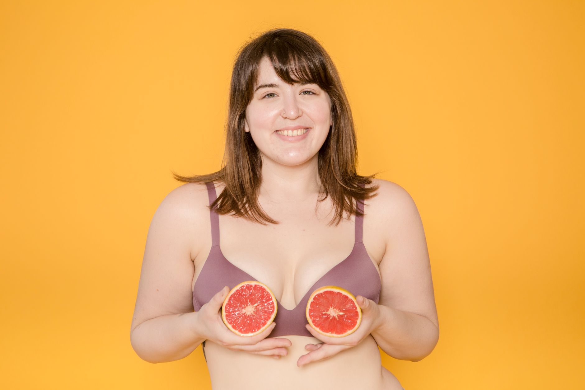 Cheerful plus size ethnic woman in bra with fresh grapefruit halves looking at camera on yellow background