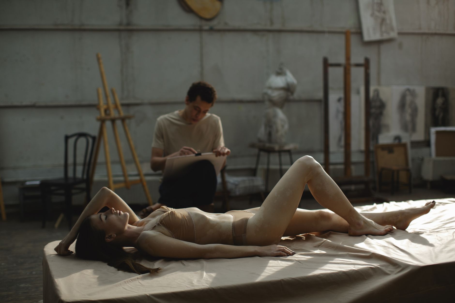 Artistic scene of a model posing nude in a studio with an artist sketching in the background.