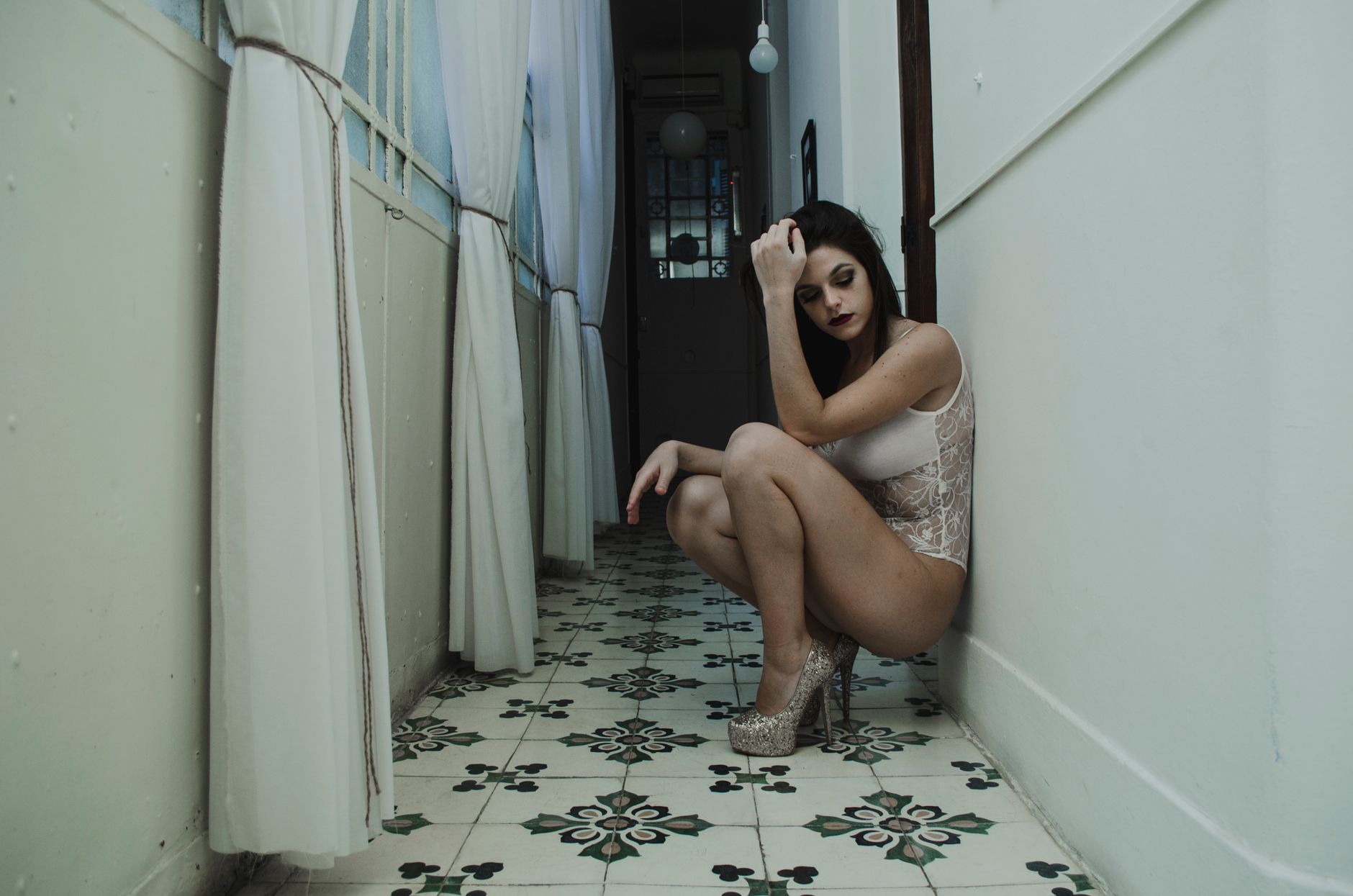 A woman poses thoughtfully in a hallway, wearing delicate lingerie and high heels.