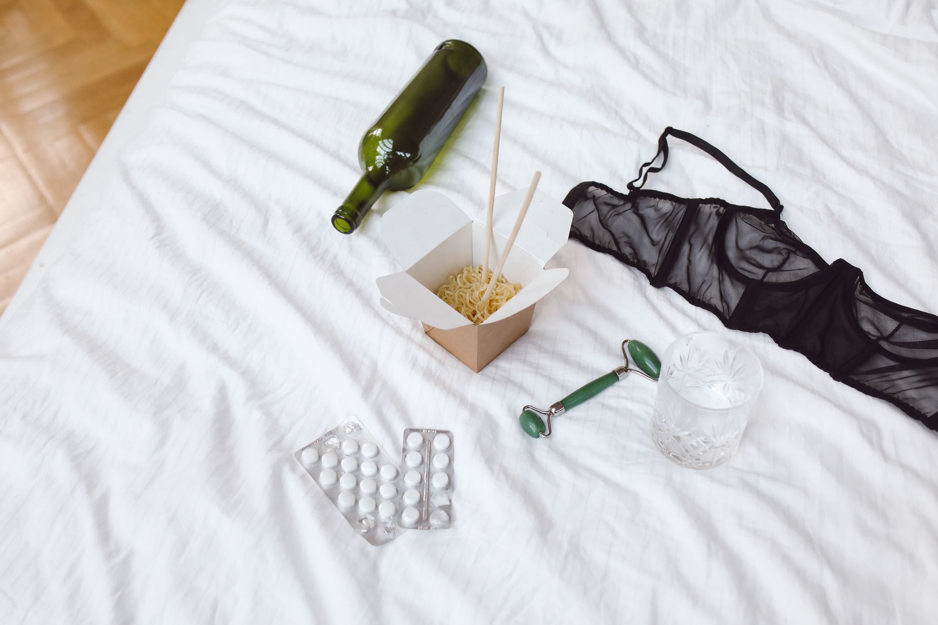A still life with afterparty items on white bedsheets, including lingerie and wine.