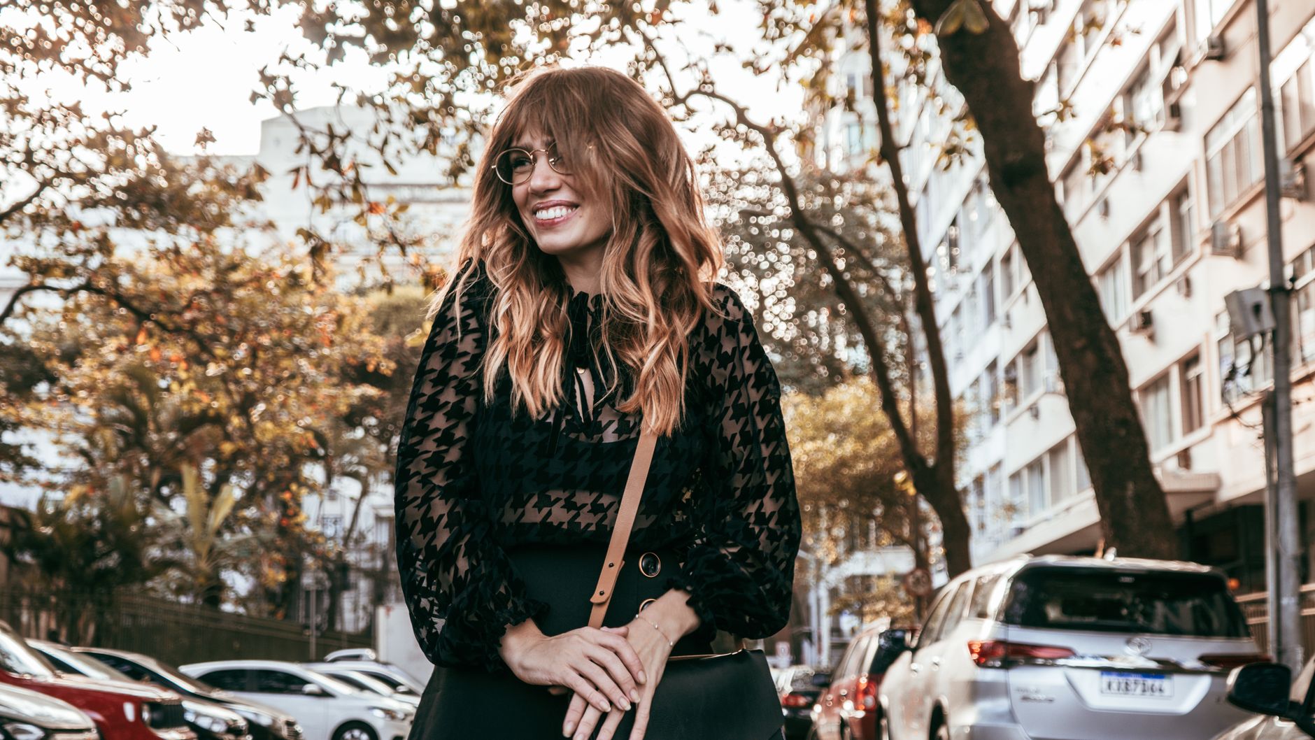 Fashionable woman with long hair and glasses smiling in a city street scene with cars.