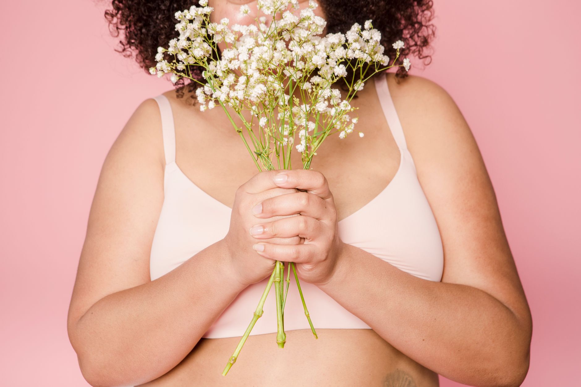 Unrecognizable plus size African American female model in lingerie standing on pink background with small flowers in hands in studio
