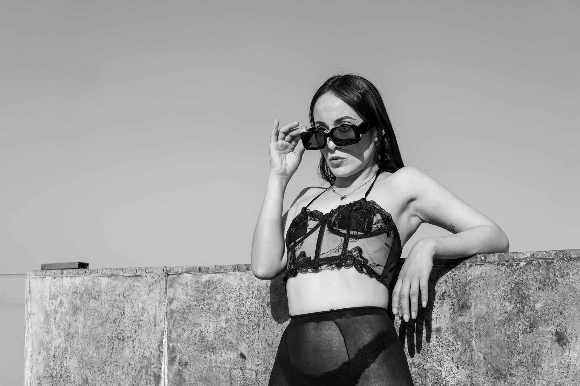 Stylish woman in lingerie and sunglasses posing against a concrete wall in black and white.