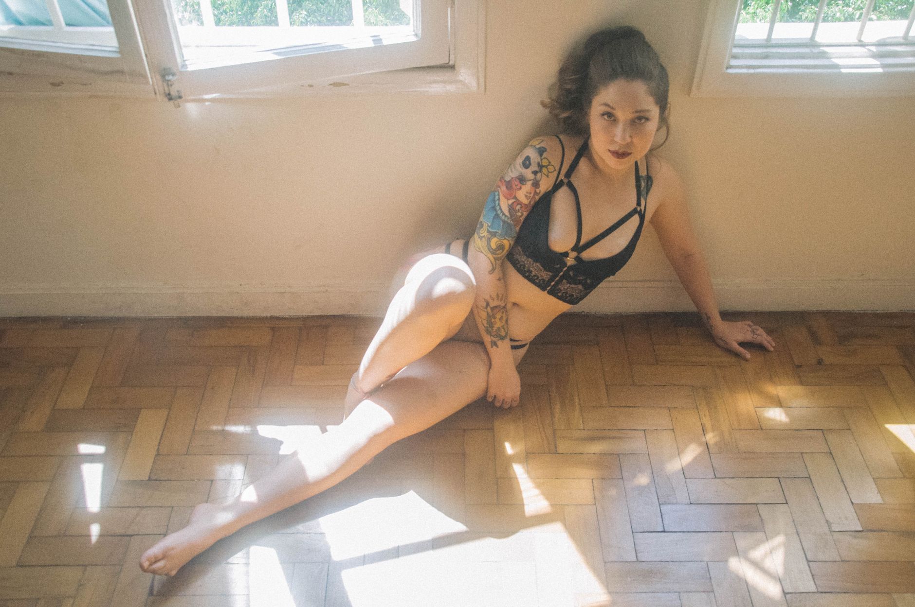 Model sitting in sunlight by a window, wearing lingerie and showcasing tattoos.