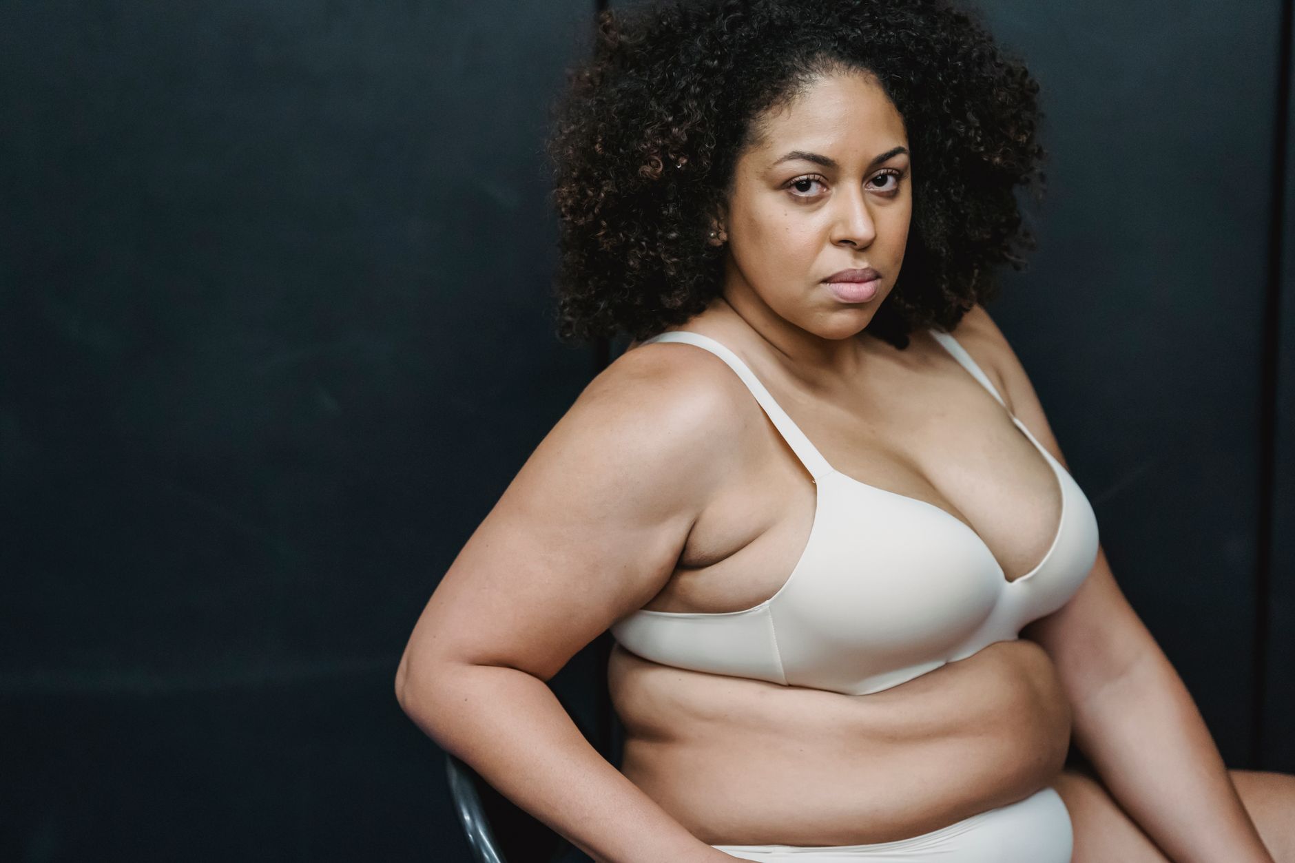 Pensive adult ethnic plump woman in white underwear sitting on chair in light studio on gray background and looking at camera