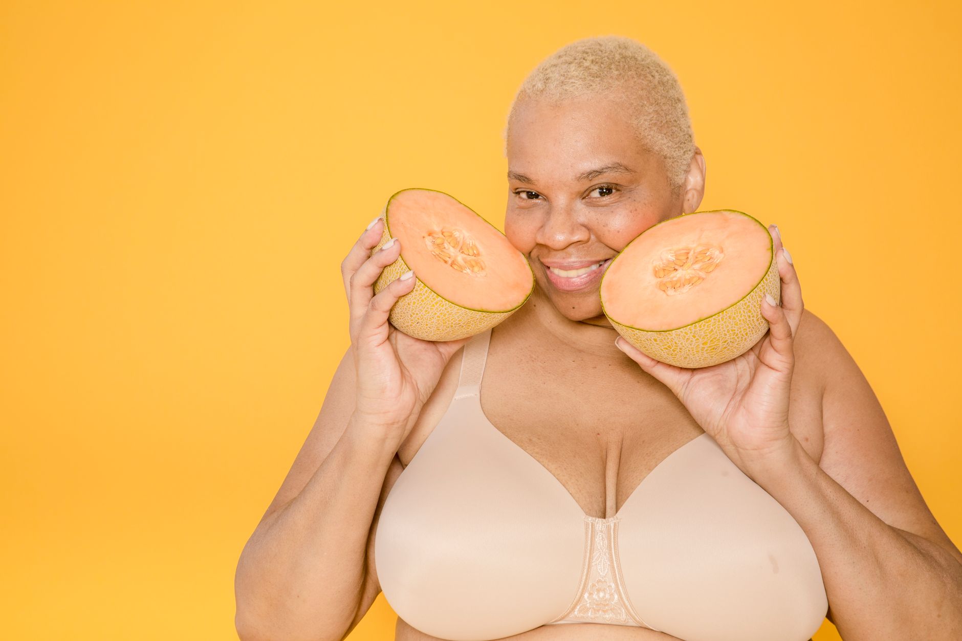 Smiling plump ethnic female in bra with fresh melon halves looking at camera on yellow background