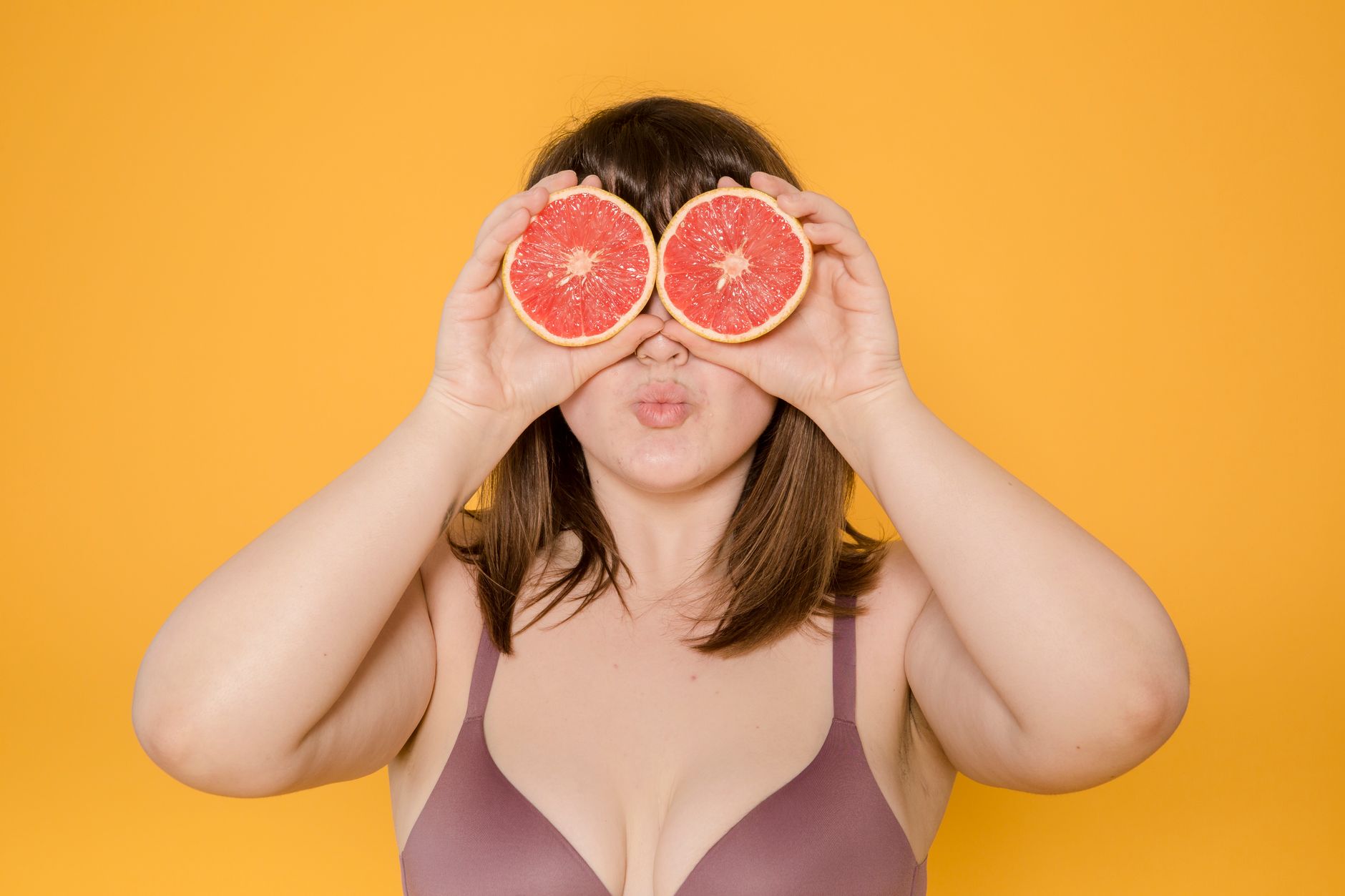 Unrecognizable overweight female wearing lingerie covering eyes with halves of grapefruit while standing on yellow background in light modern studio