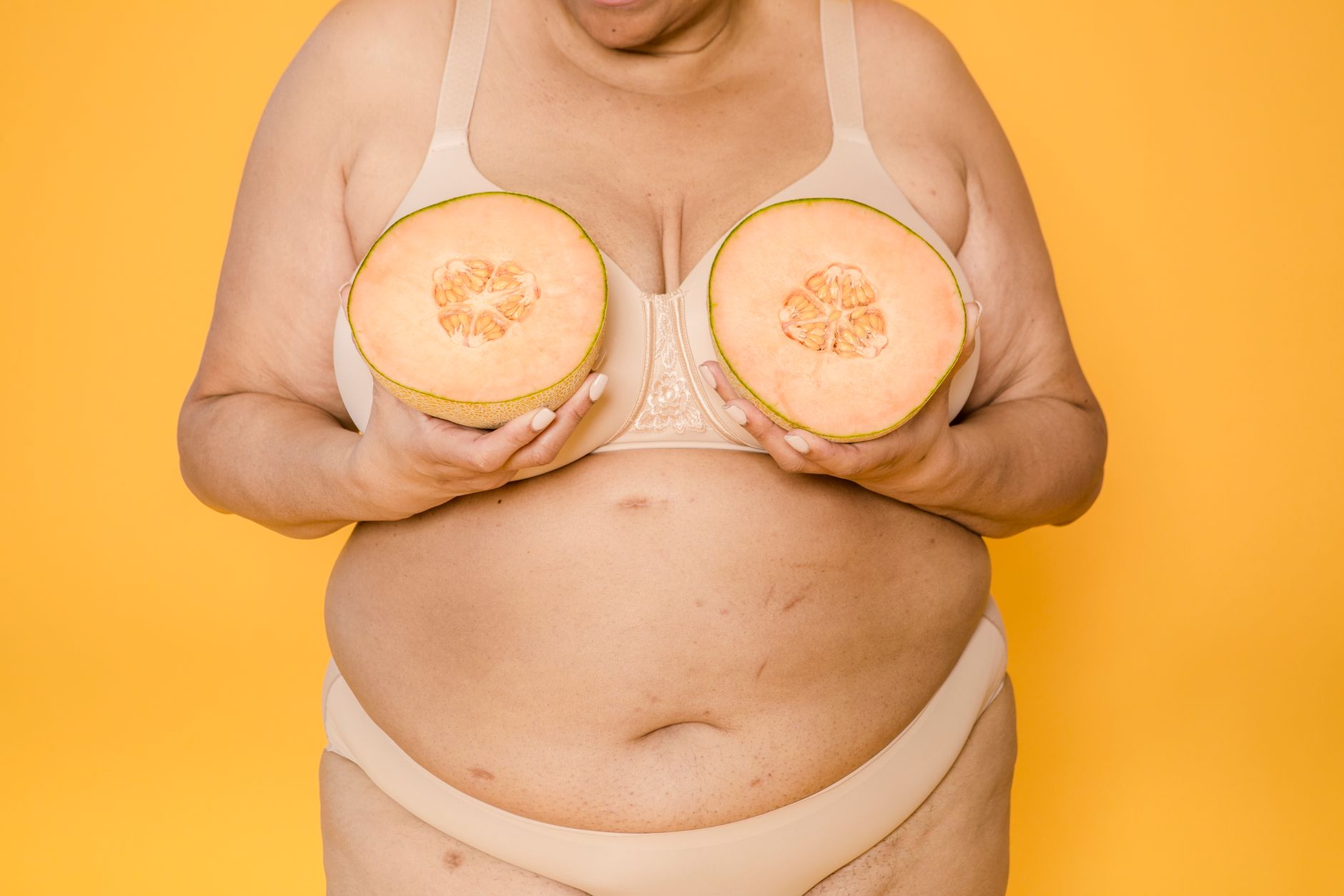 Crop unrecognizable voluptuous female model in bikini and brassiere with fresh melon halves on yellow background
