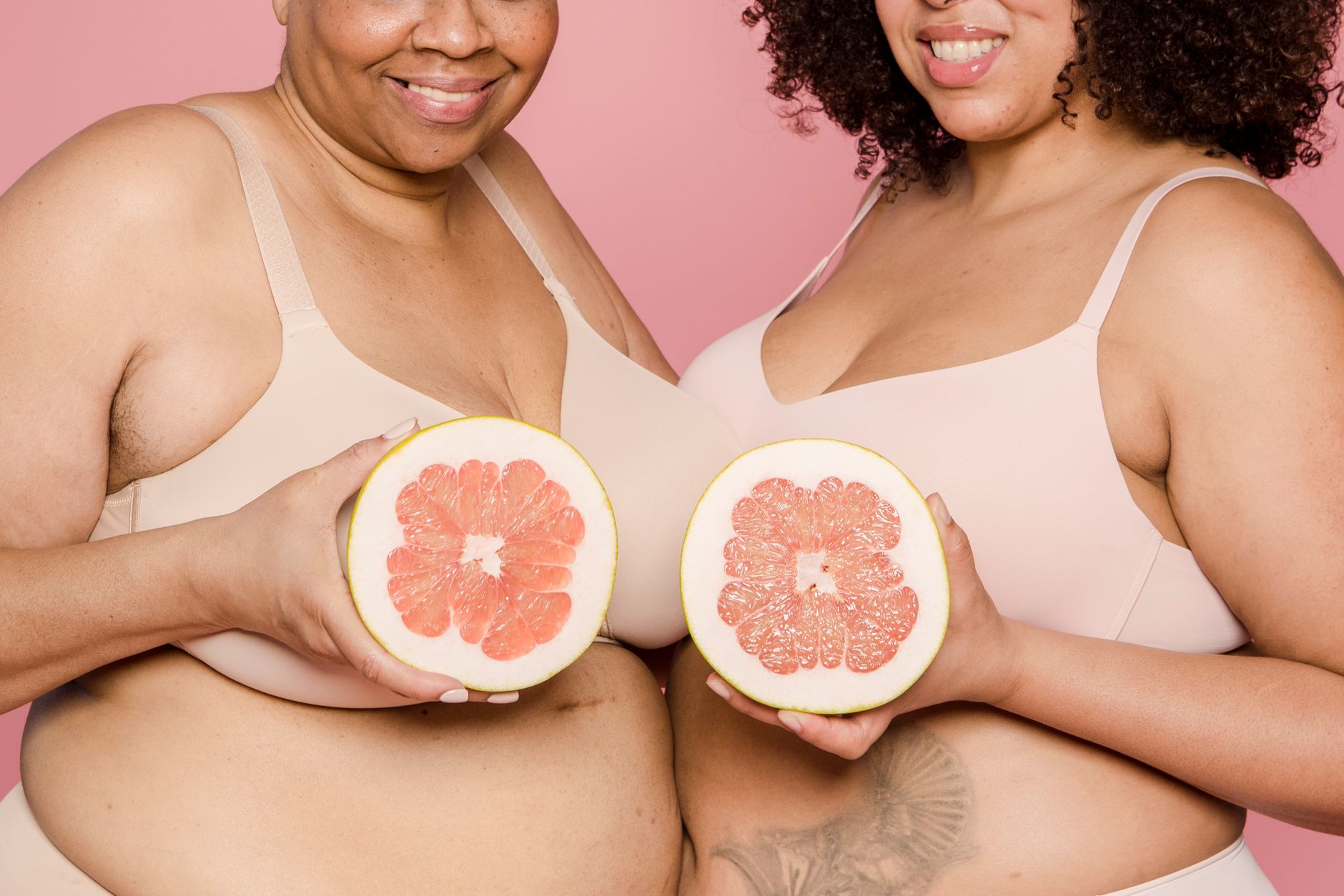 Anonymous plus size African American female models in lingerie with halves of ripe grapefruit in hands smiling on pink background in studio