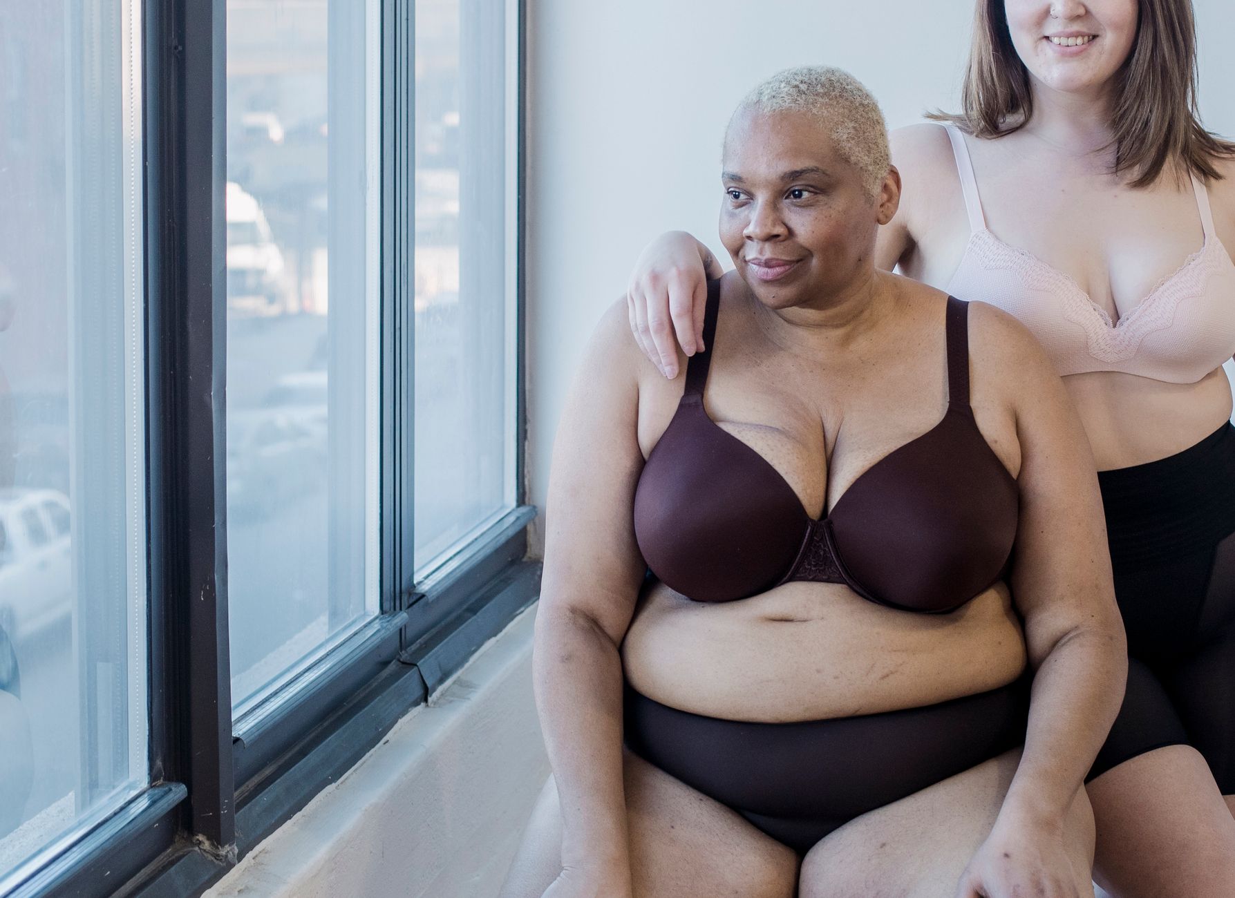 Crop plus size female and overweight African American woman in lingerie spending time together in light room near big window