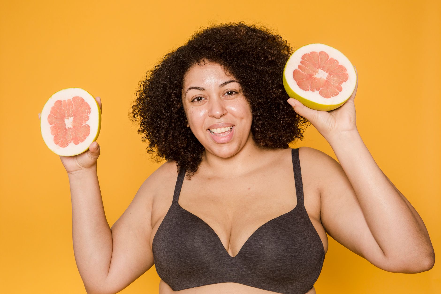 Cheerful plus size African American female in bra with curly hair and cut juicy grapefruit looking at camera on yellow background